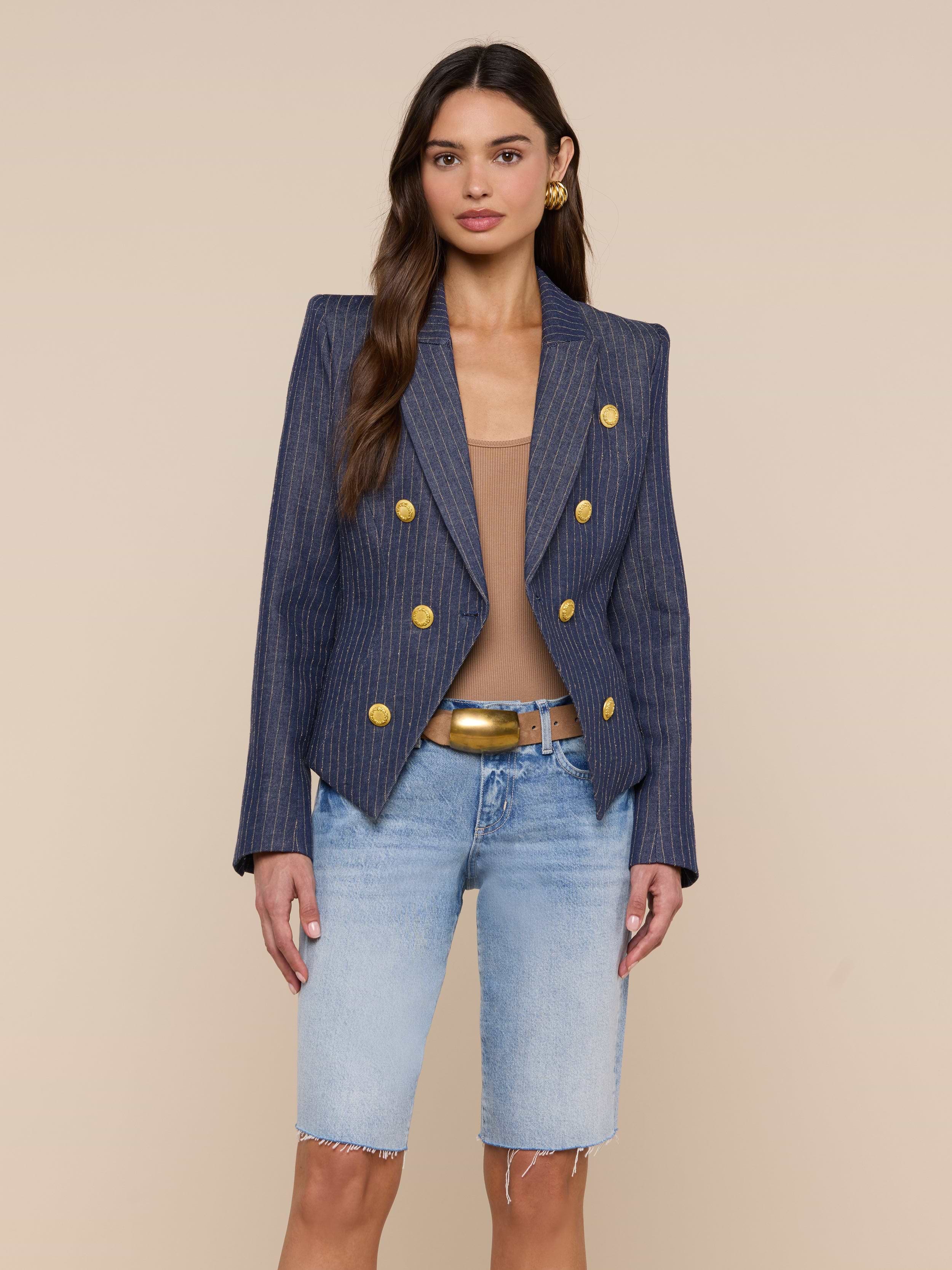 A woman wears the Solar Pinstriped Blazer—a slim fit, linen piece with gold buttons—paired with a tan top, denim Bermuda shorts, and a tan belt. She stands against a plain light background, looking at the camera.