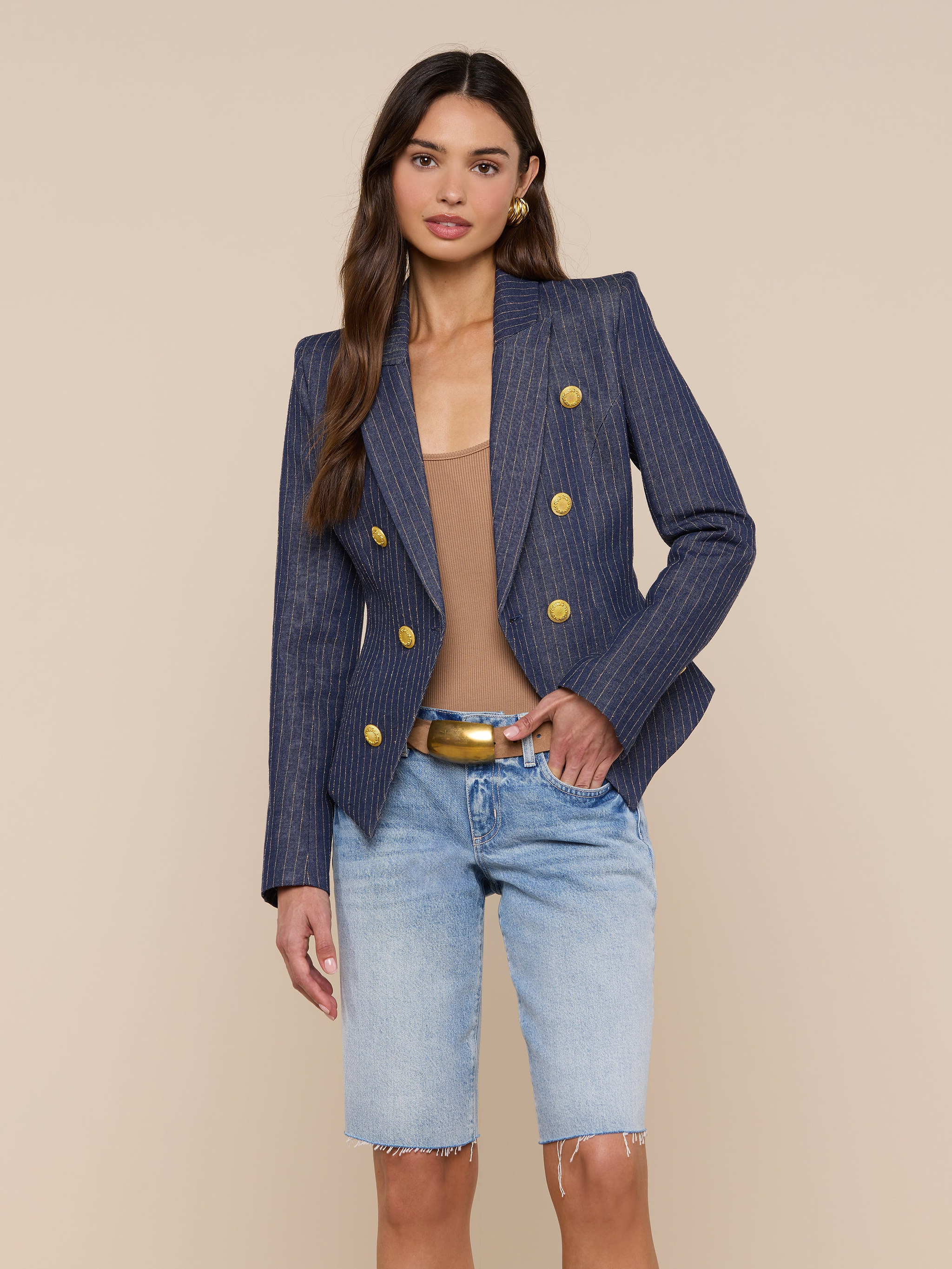 A woman stands against a plain light background wearing the Solar Pinstriped Blazer—a navy double-breasted linen blazer with gold buttons—paired with a beige top, tan belt, and light blue knee-length denim shorts.