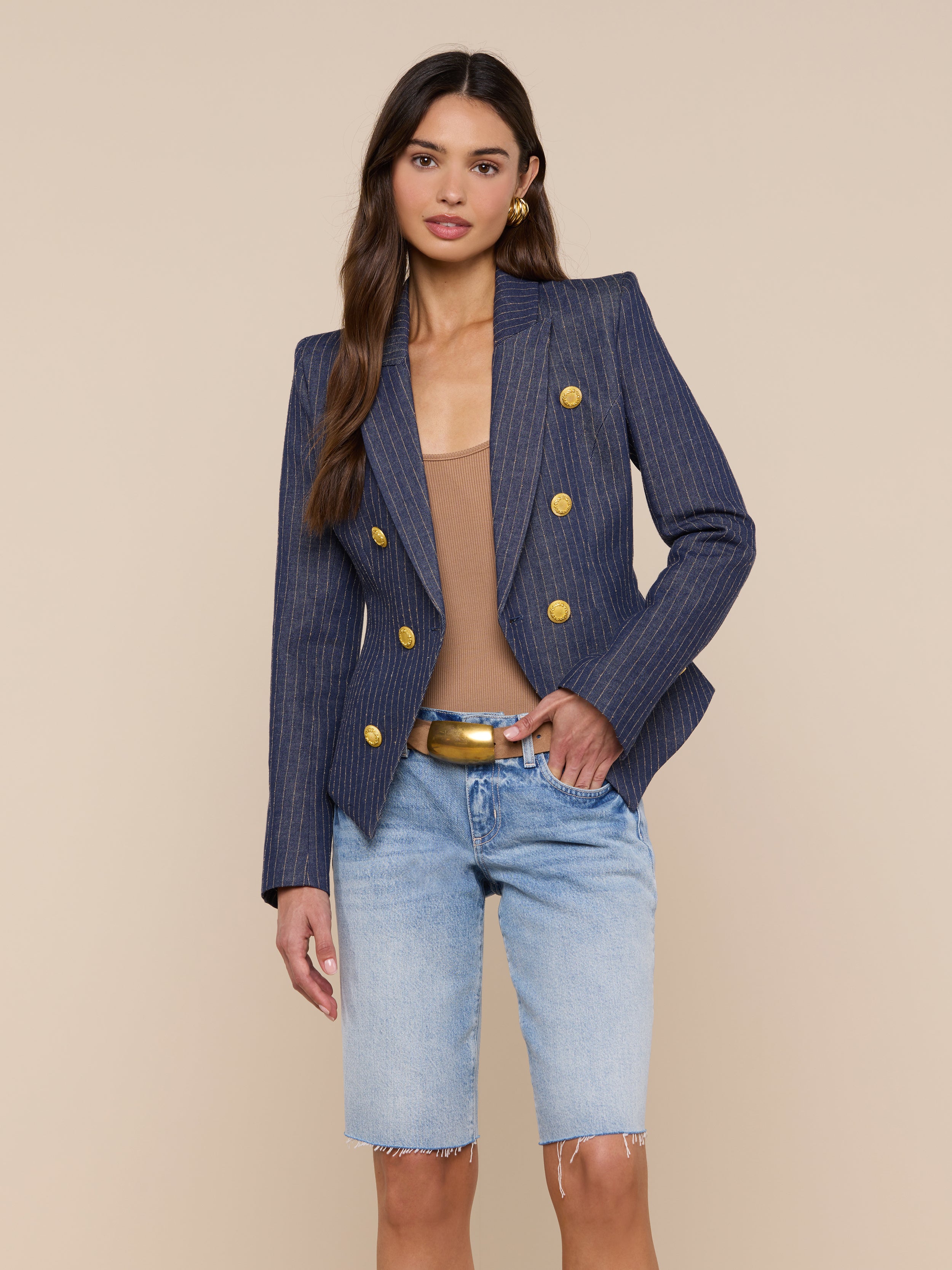 A woman stands against a plain light background wearing the Solar Pinstriped Blazer—a navy double-breasted linen blazer with gold buttons—paired with a beige top, tan belt, and light blue knee-length denim shorts.