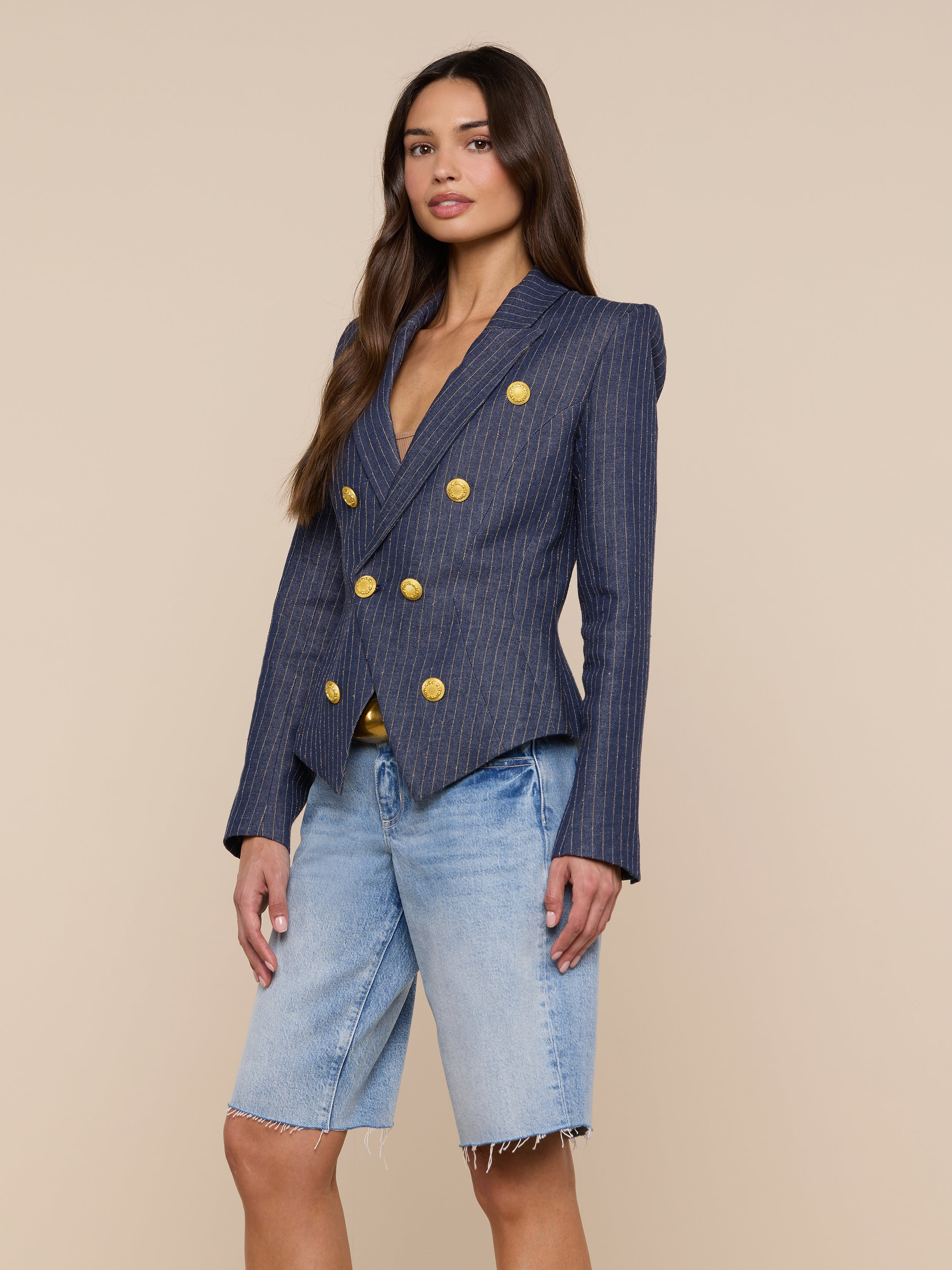 A woman poses against a plain light background wearing the Solar Pinstriped Blazer—navy linen with gold buttons—paired with light blue knee-length denim shorts.