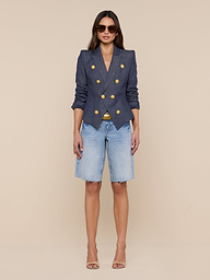 A woman stands against a white background wearing sunglasses, the Solar Pinstriped Blazer featuring gold buttons, light blue knee-length denim shorts, and nude high-heeled sandals.