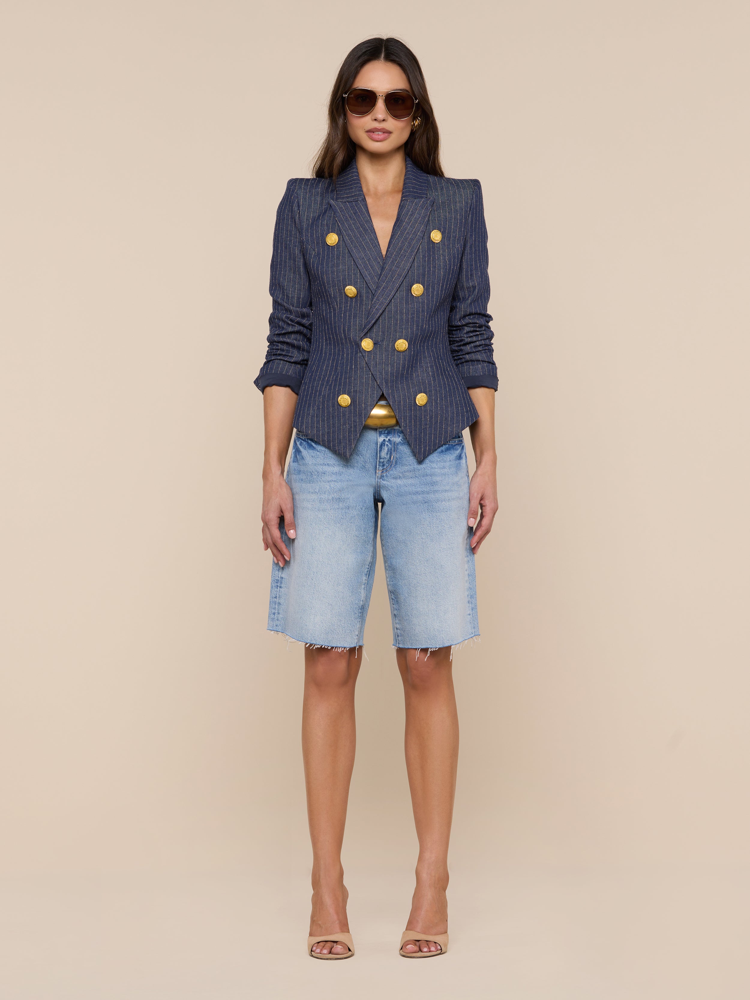 A woman stands against a white background wearing sunglasses, the Solar Pinstriped Blazer featuring gold buttons, light blue knee-length denim shorts, and nude high-heeled sandals.