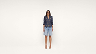 Woman wearing sunglasses, a navy double-breasted blazer with gold buttons, denim knee-length shorts, and nude high heels stands against a plain white background.