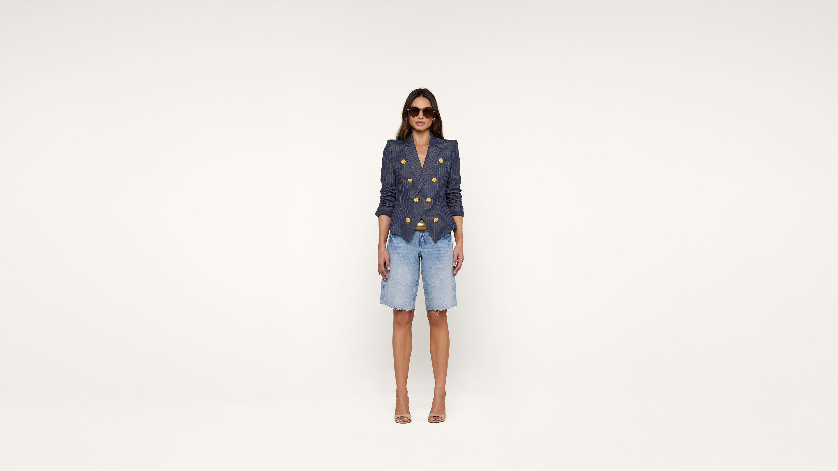 Woman wearing sunglasses, a navy double-breasted blazer with gold buttons, denim knee-length shorts, and nude high heels stands against a plain white background.