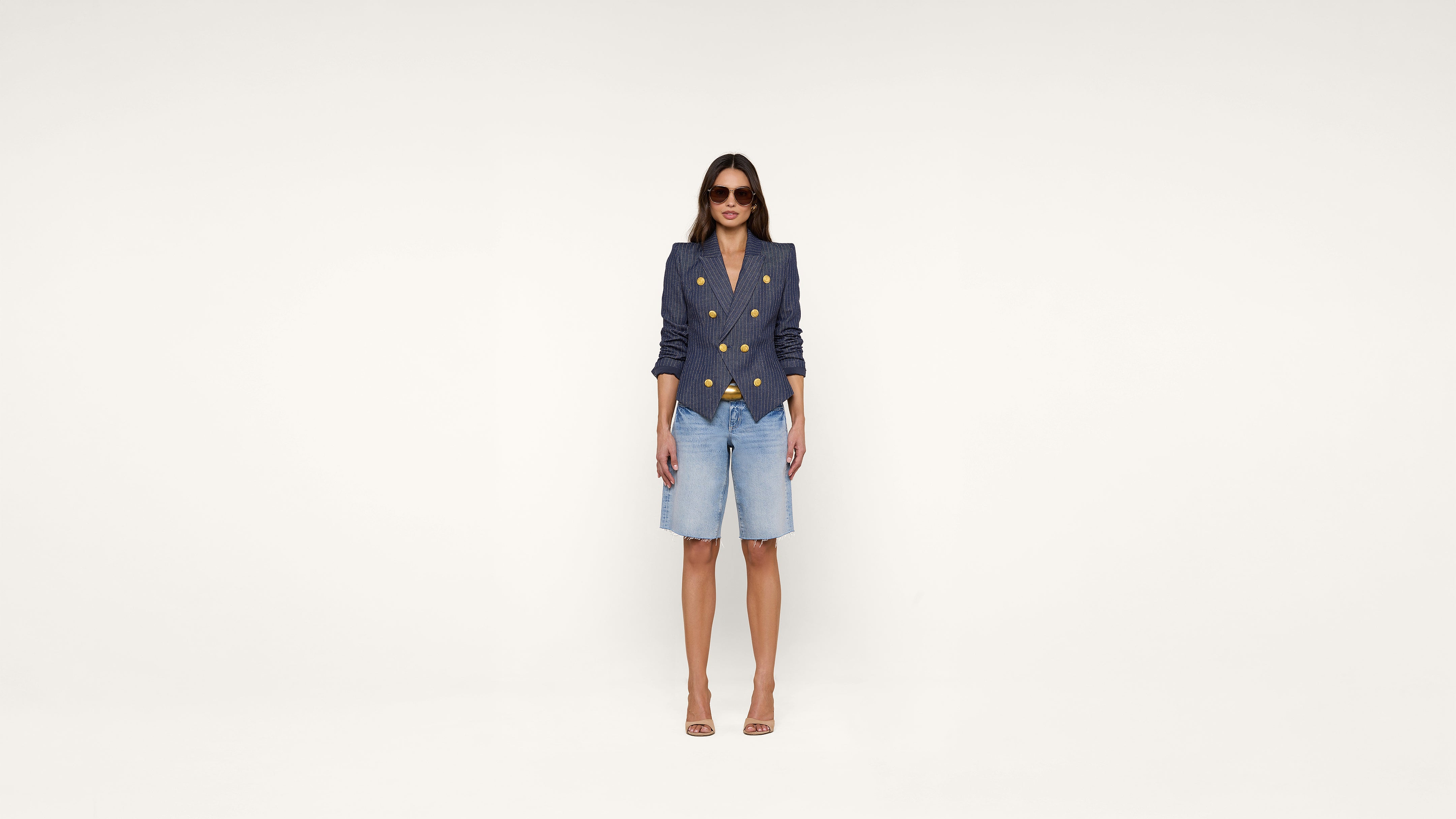 Woman wearing sunglasses, a navy double-breasted blazer with gold buttons, denim knee-length shorts, and nude high heels stands against a plain white background.