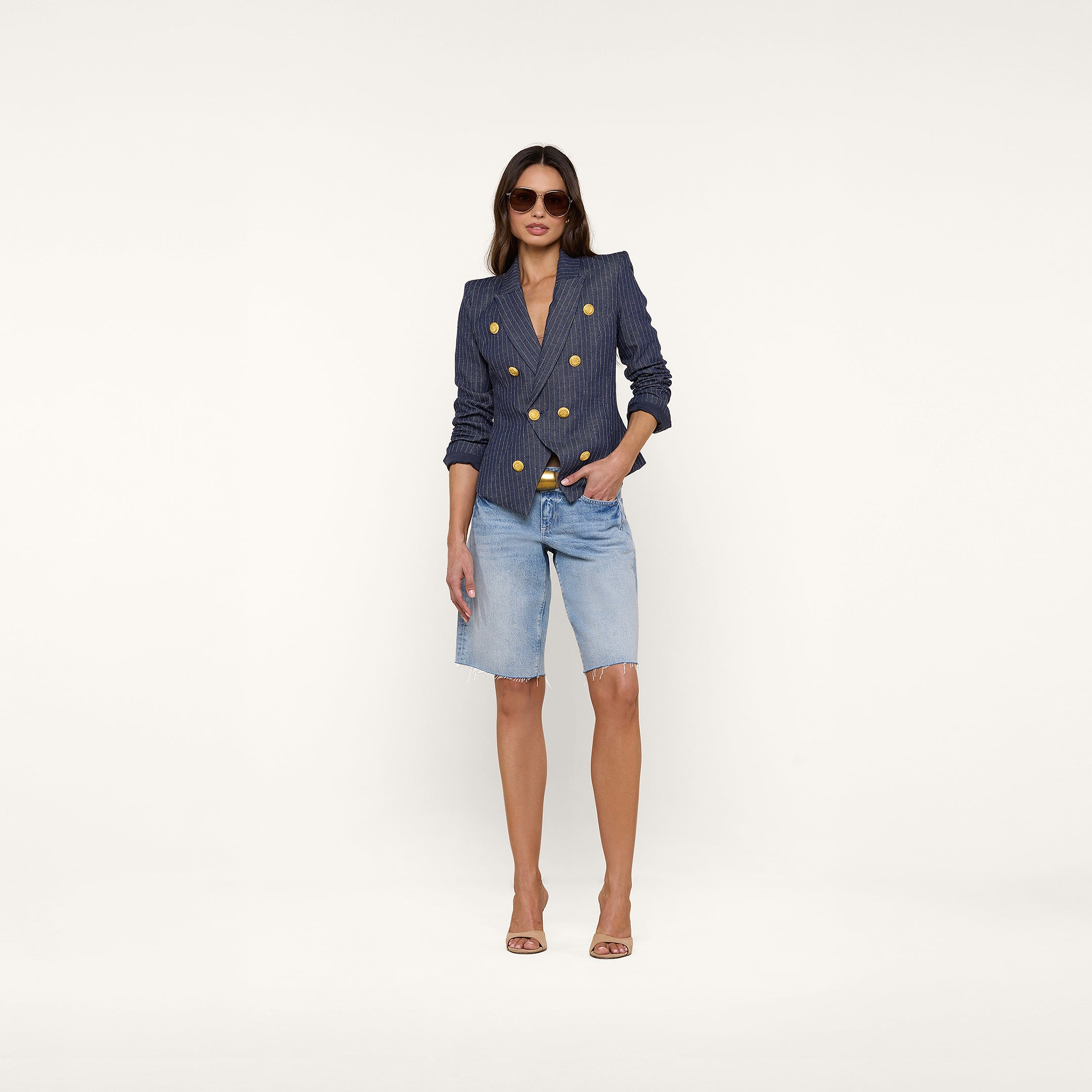 A woman wearing the Solar Pinstriped Blazer—a navy slim-fit linen blazer—styled with denim knee-length shorts, nude heels, and sunglasses stands against a plain white background.