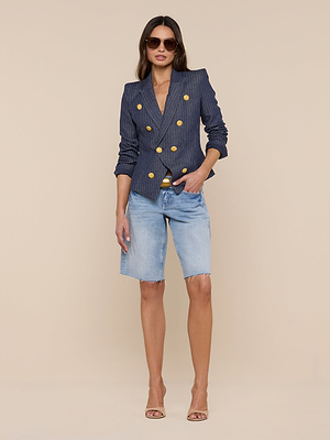 A woman in sunglasses and the Solar Pinstriped Blazer—navy blue, slim fit, gold buttons—paired with light blue denim Bermuda shorts and nude heeled sandals stands against a plain beige background.