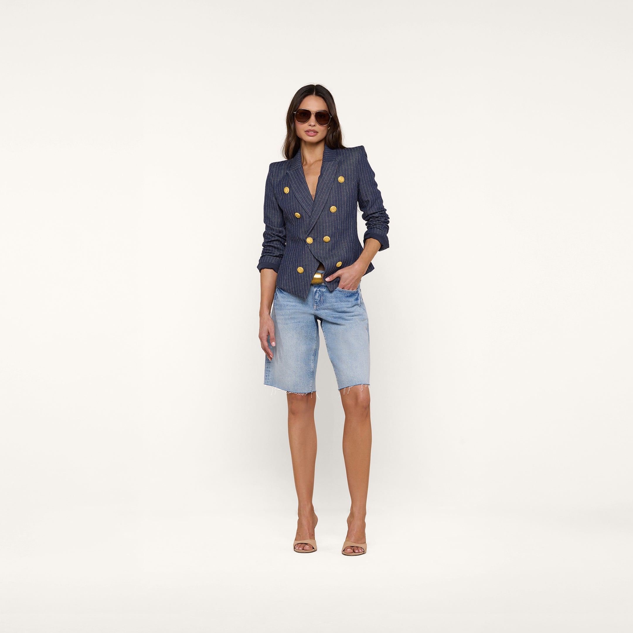 A woman wearing the Solar Pinstriped Blazer—a navy slim-fit linen blazer—styled with denim knee-length shorts, nude heels, and sunglasses stands against a plain white background.