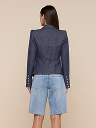 A woman stands facing away, wearing the Solar Pinstriped Blazer— a double-breasted linen piece featuring gold buttons on the sleeves—paired with knee-length, light blue denim shorts. The background is plain and neutral.