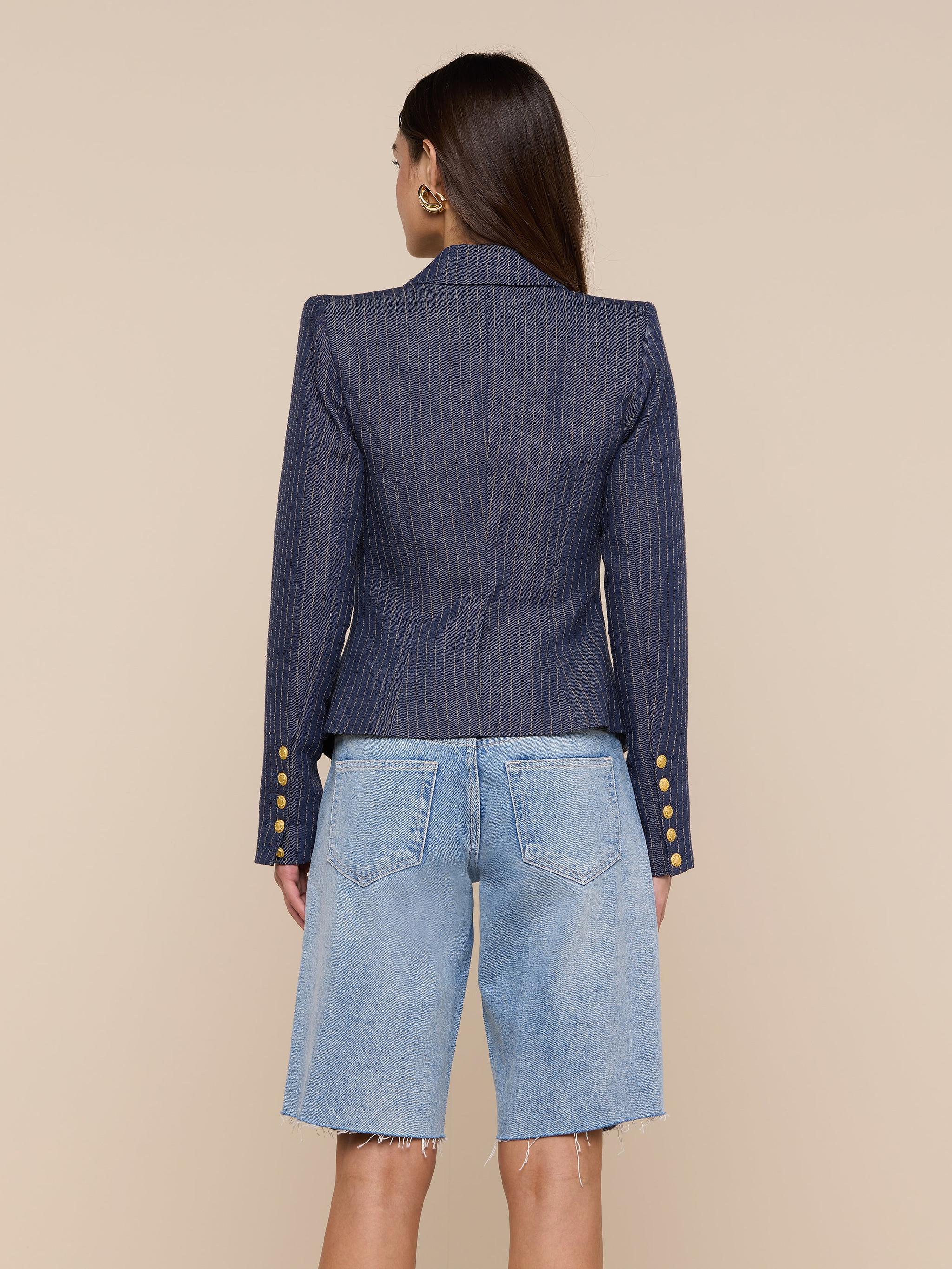 A woman stands facing away, wearing the Solar Pinstriped Blazer— a double-breasted linen piece featuring gold buttons on the sleeves—paired with knee-length, light blue denim shorts. The background is plain and neutral.