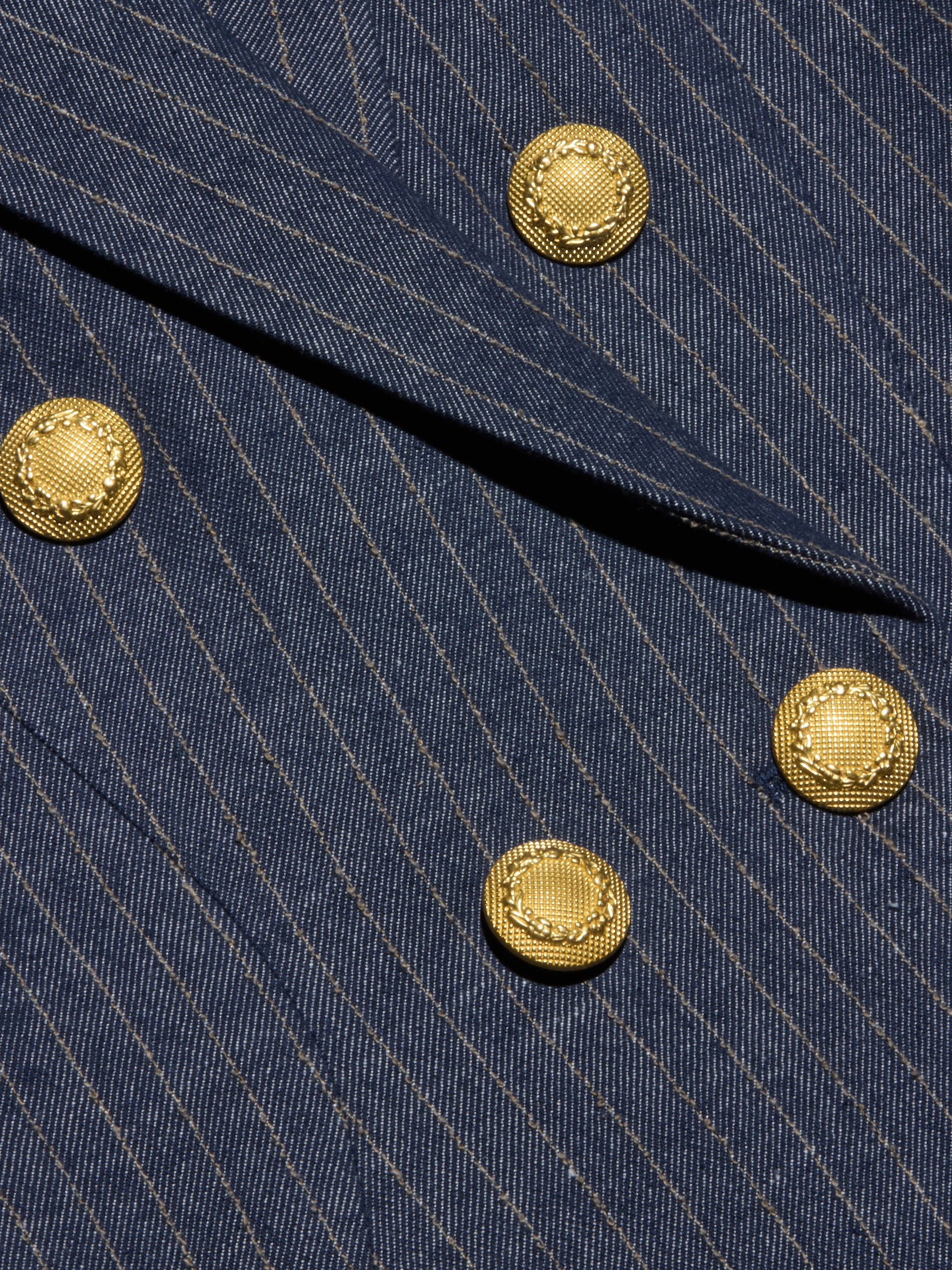 Close-up of the Solar Pinstriped Blazer in blue, showcasing its pinstripe fabric, folded lapel, and four decorative gold buttons—suggesting a tailored double-breasted design.