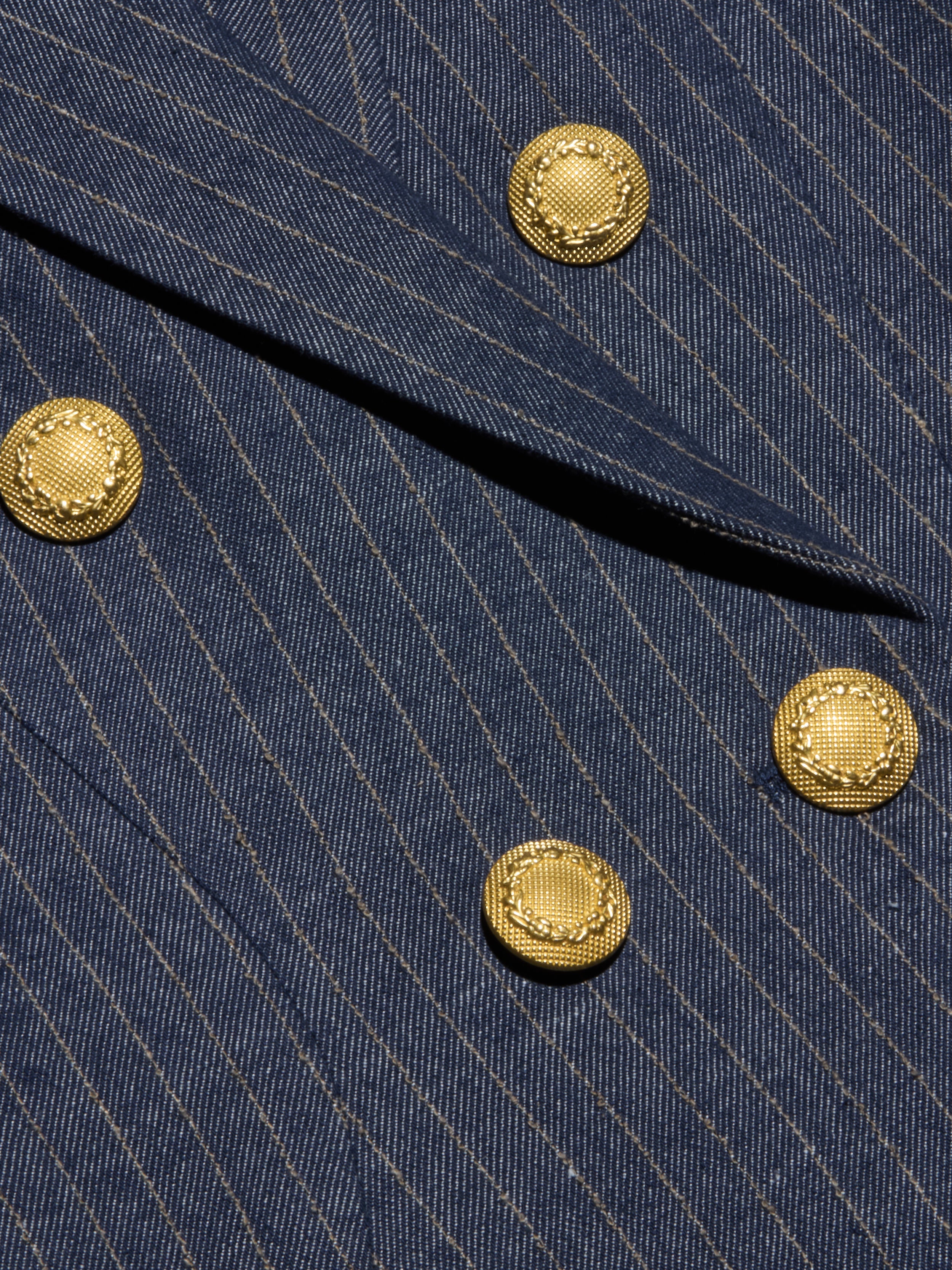 Close-up of the Solar Pinstriped Blazer in blue, showcasing its pinstripe fabric, folded lapel, and four decorative gold buttons—suggesting a tailored double-breasted design.
