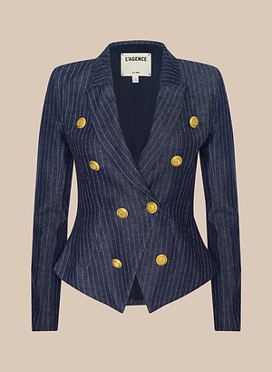 The Solar Pinstriped Blazer features a fitted navy linen silhouette, double-breasted front, wide lapels, and eight gold buttons. L’AGENCE is labeled inside the collar. Set against a beige background.