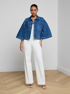 A woman stands indoors on a wooden floor, facing forward against a plain light wall. She wears the Silas Cape Jacket in all-cotton denim with cape sleeves, styled over a white top, high-waisted white pants, and white heeled sandals.