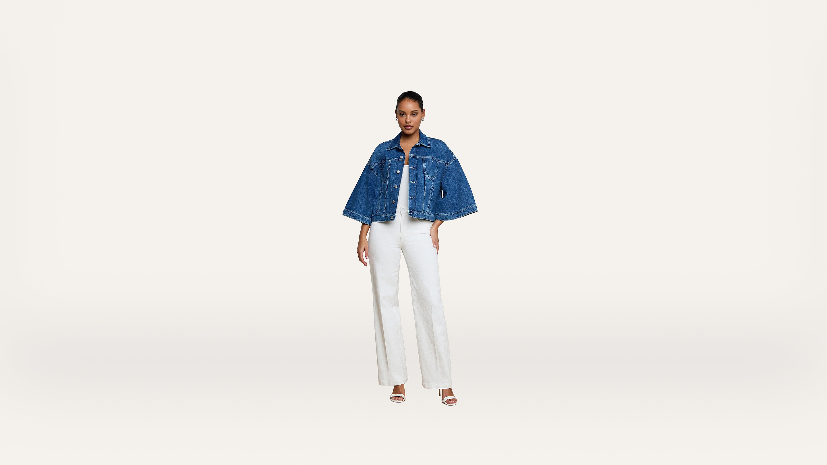 Woman stands facing forward, wearing a blue denim jacket over a white top and white wide-leg pants, with white heeled sandals, on a plain white background.