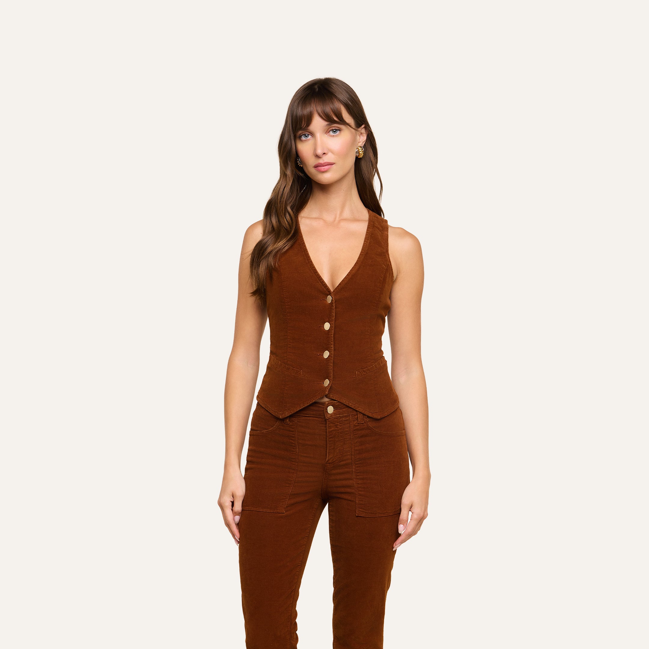 A woman with long brown hair and bangs stands against a plain background, wearing the Yelena Vest in brown stretch corduroy paired with matching brown pants. Her expression is neutral.