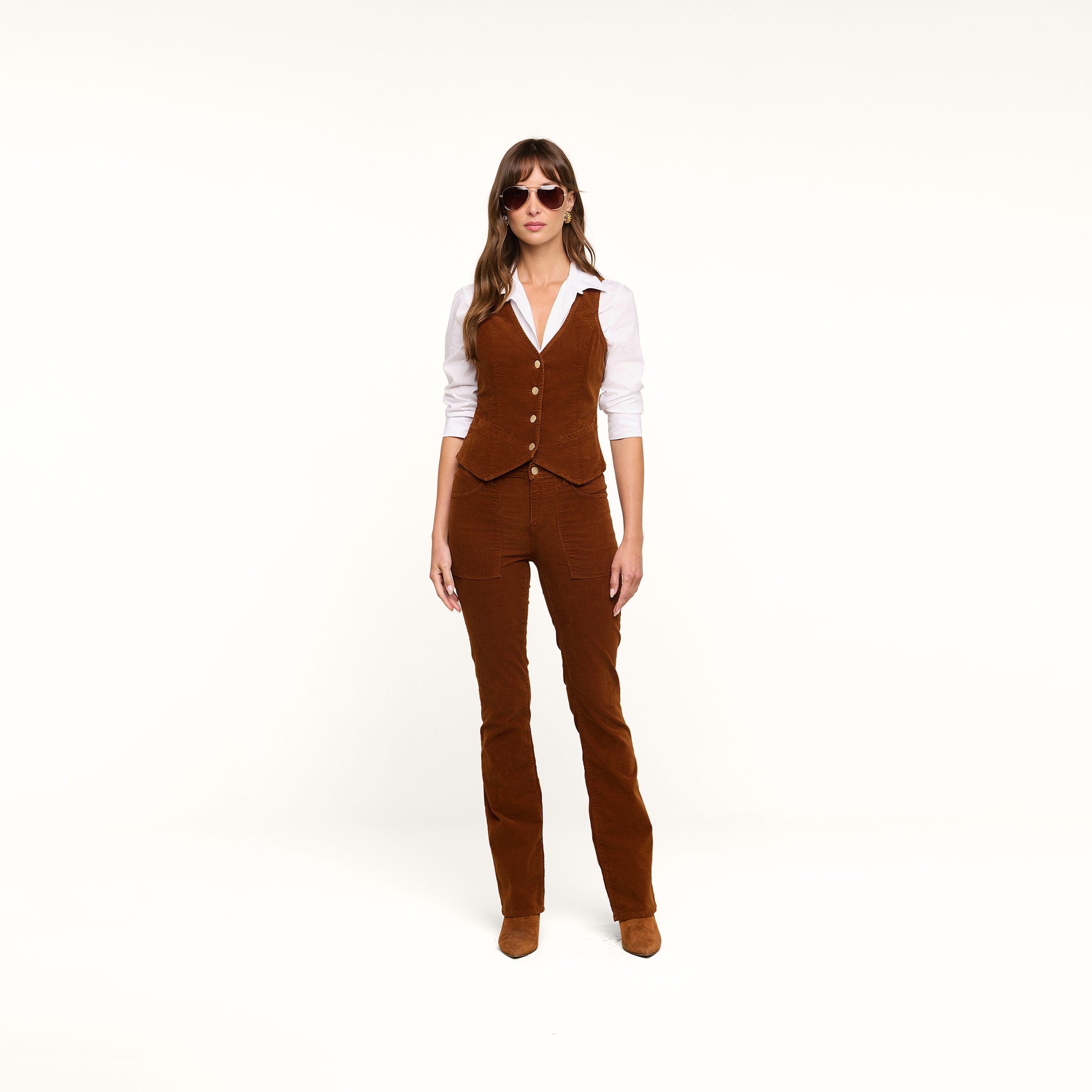 A woman stands facing forward on a plain white background, arms at her sides, wearing sunglasses, a white shirt, the Yelena Vest, and matching casual corduroy brown pants.