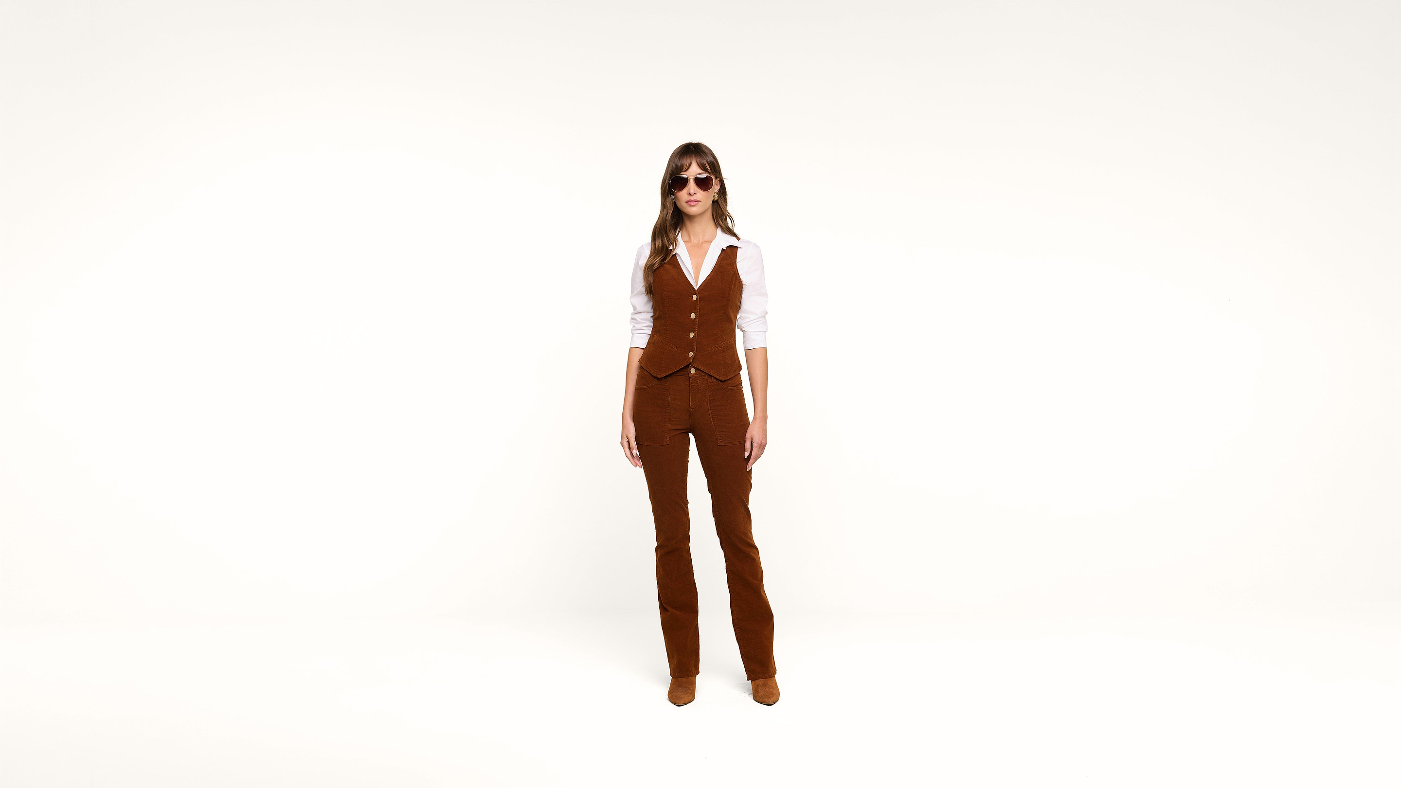 A woman stands against a plain white background, wearing sunglasses, a white shirt, and a brown vest with matching brown flared pants and shoes.