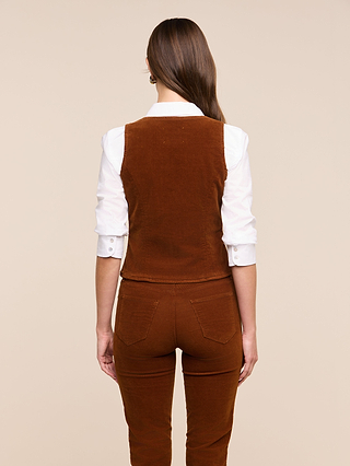 A woman with long brown hair stands facing away, wearing a white long-sleeve shirt layered under the Yelena Vest and matching brown stretch corduroy pants, set against a plain beige background.