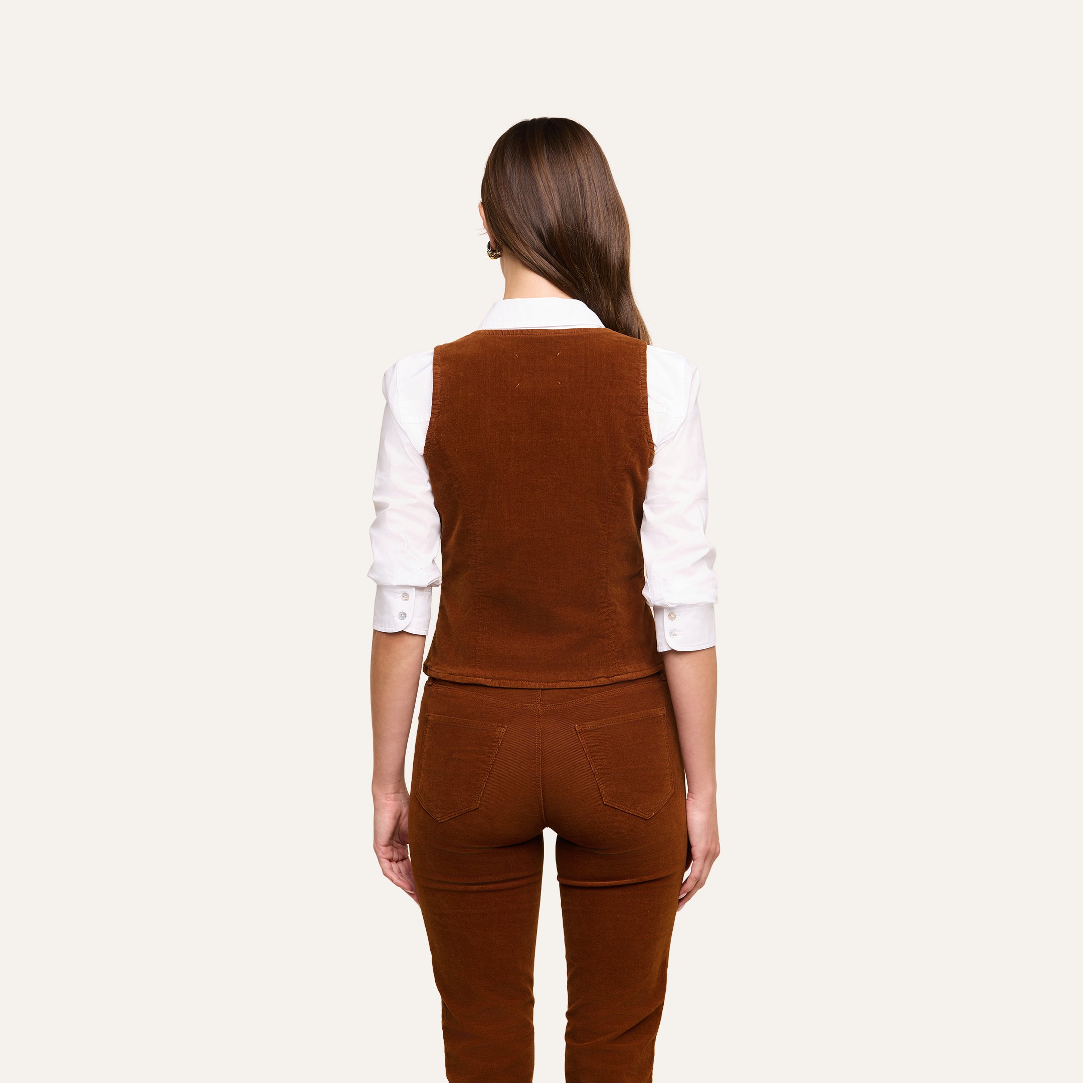Wearing the Yelena Vest over a white long-sleeve shirt and matching stretch corduroy pants, a woman stands with her straight brown hair falling down her back against a plain light background.