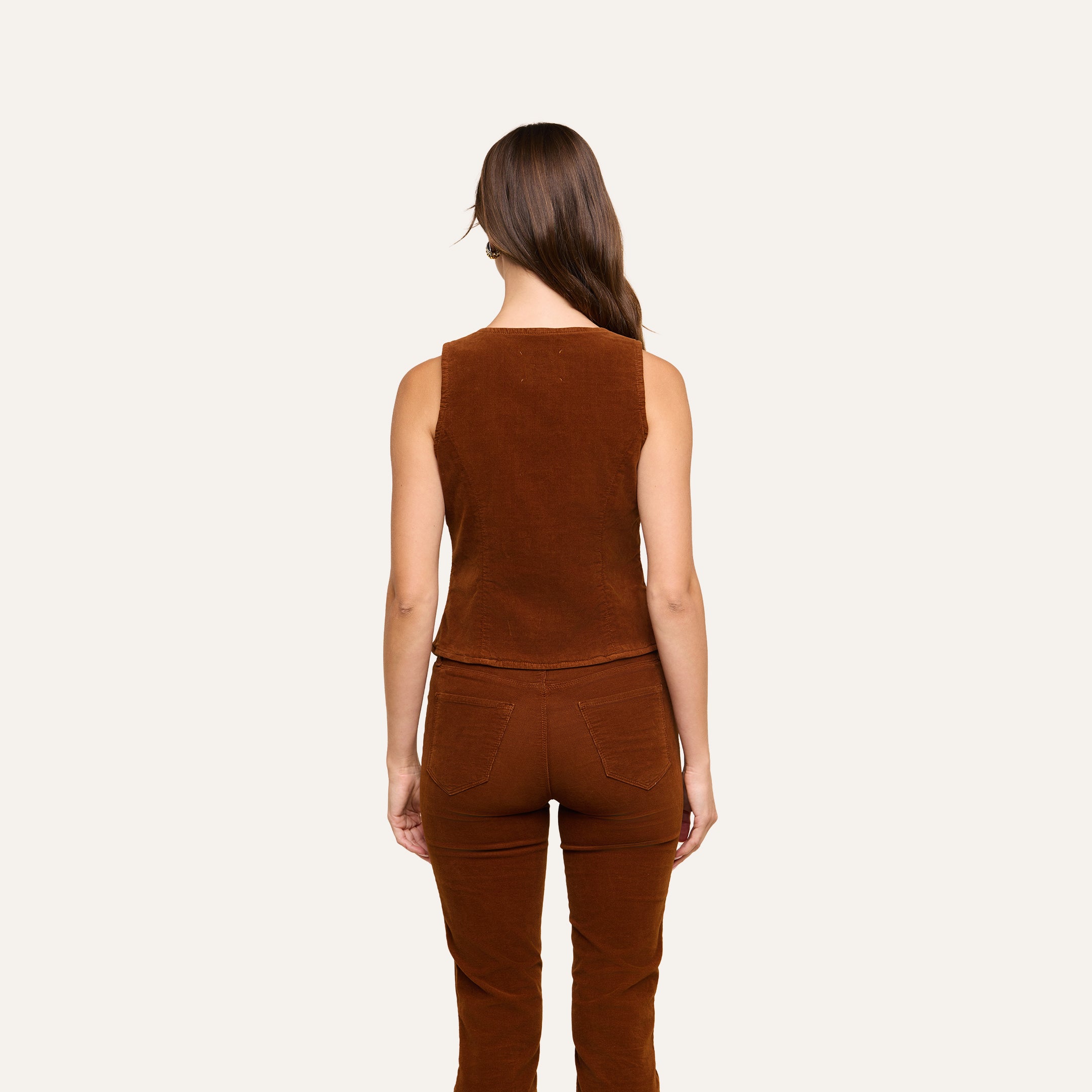 A woman with long brown hair, wearing the Yelena Vest and casual corduroy pants, stands facing away against a plain light background.