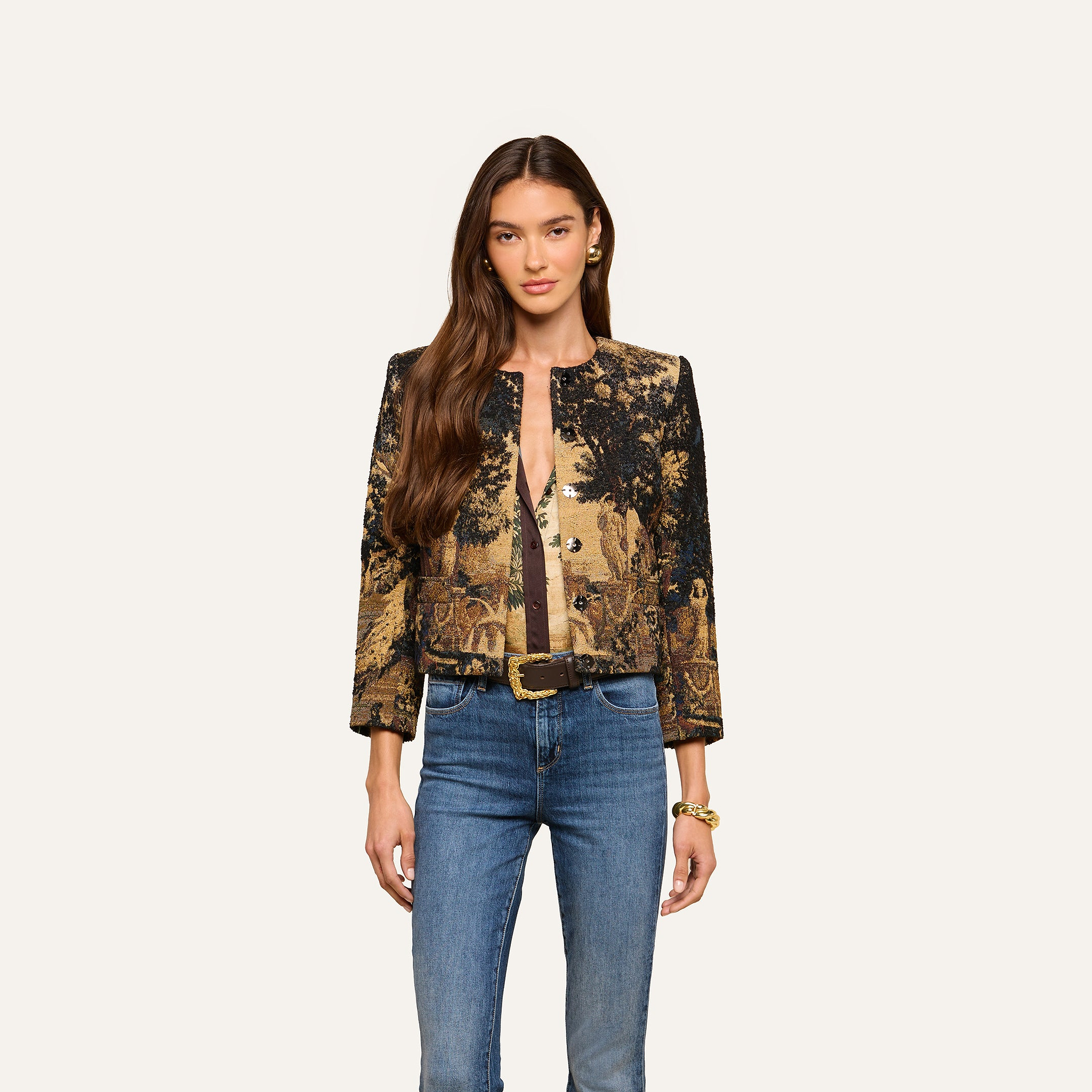 A woman with long brown hair stands against a plain light background, facing forward with a neutral expression. She is wearing the Lian Tapestry Jacket—a gold and black patterned piece reminiscent of a Flemish tapestry—paired with a brown top, blue jeans, and a gold belt.