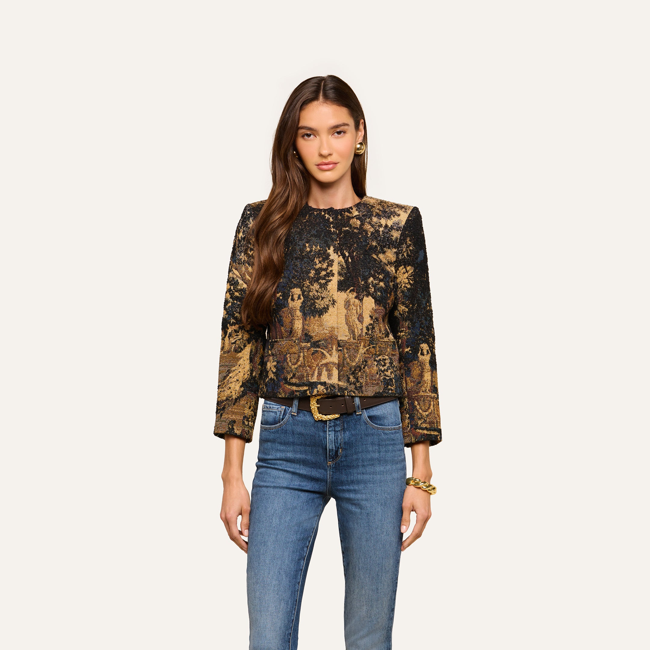 Wearing the Lian Tapestry Jacket—a black and gold patterned piece reminiscent of a Flemish tapestry—a woman with long brown hair stands against a plain light background, styled with blue jeans, a black belt, and gold jewelry.