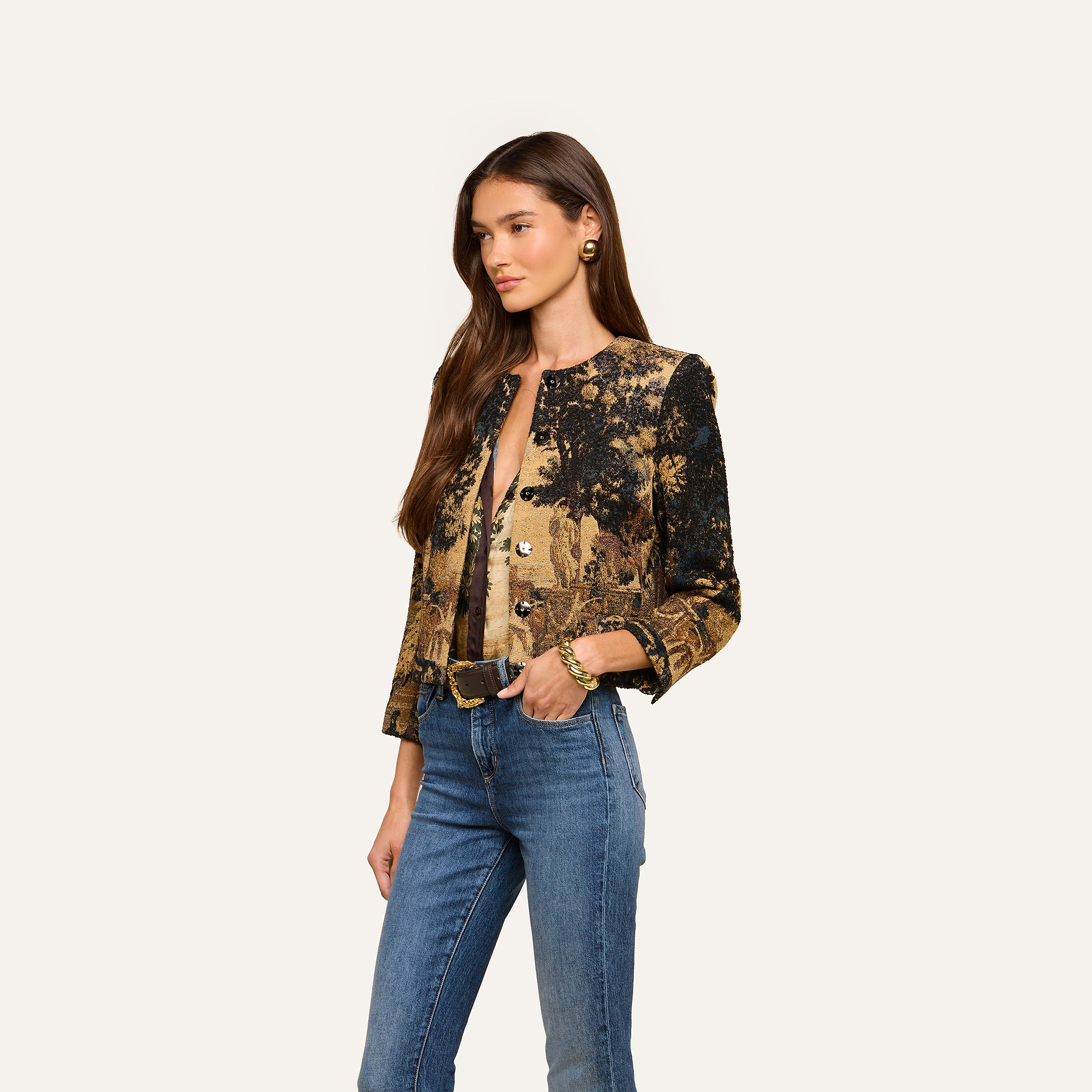 A woman with long brown hair wears the Lian Tapestry Jacket—featuring a gold and black Flemish-inspired pattern with large buttons—paired with blue jeans and gold jewelry, standing against a plain light background.