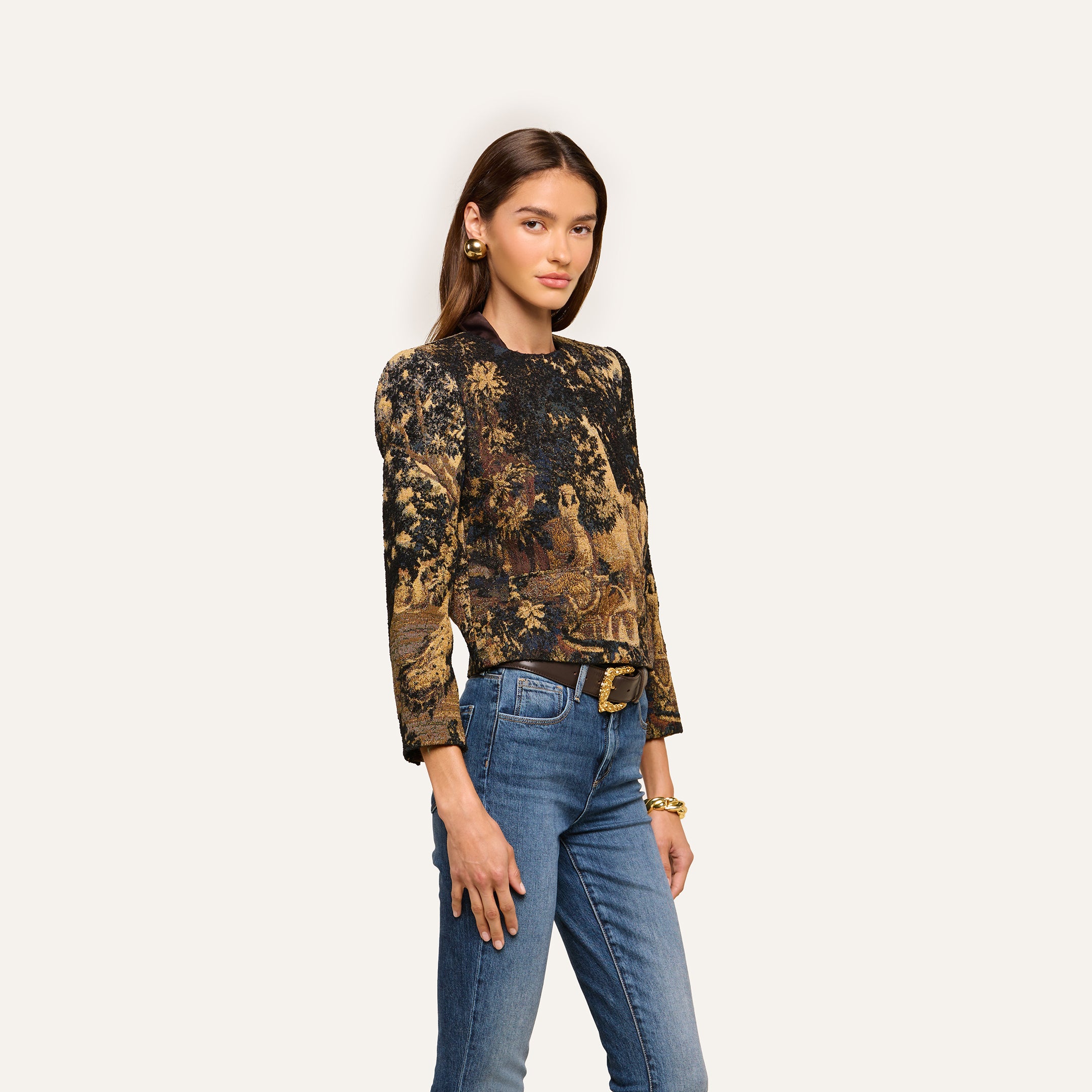 A woman stands against a plain background, wearing the Lian Tapestry Jacket—a long-sleeved piece featuring gold and black patterns inspired by Flemish tapestry. She pairs it with blue jeans, a black belt, gold earrings, and bracelets. Her long brown hair frames her neutral expression.