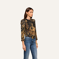 A woman with long brown hair stands against a plain light background, wearing the Lian Tapestry Jacket—a printed, long-sleeve jacket featuring intricate patterns reminiscent of Flemish tapestry—paired with blue jeans, a black belt, gold earrings, and a bracelet.