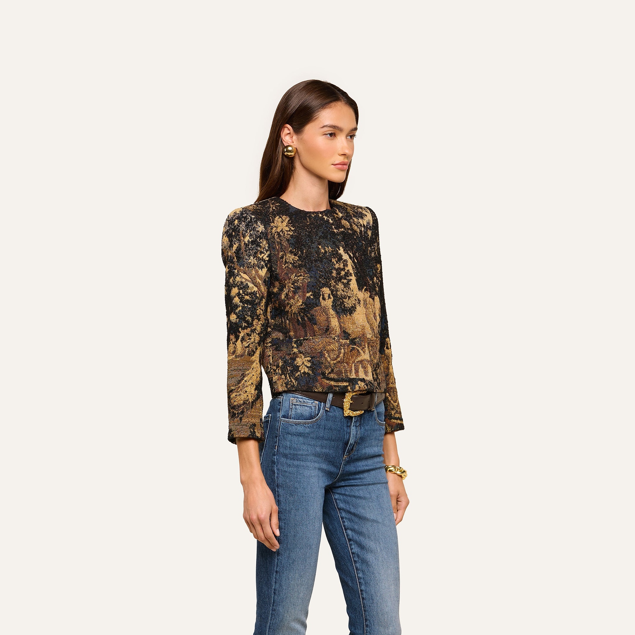 A woman with long brown hair stands against a plain light background, wearing the Lian Tapestry Jacket—a printed, long-sleeve jacket featuring intricate patterns reminiscent of Flemish tapestry—paired with blue jeans, a black belt, gold earrings, and a bracelet.