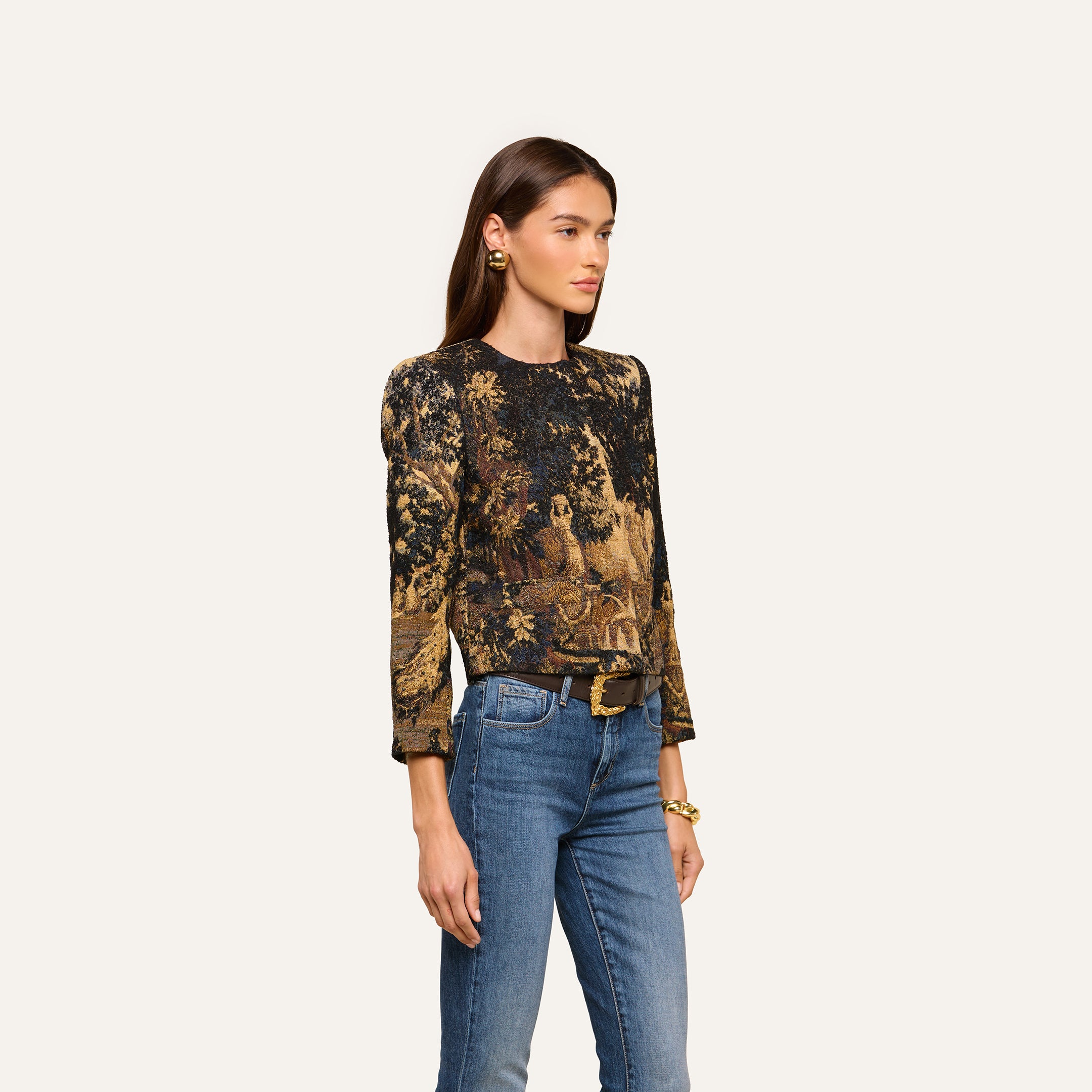A woman with long brown hair stands against a plain light background, wearing the Lian Tapestry Jacket—a printed, long-sleeve jacket featuring intricate patterns reminiscent of Flemish tapestry—paired with blue jeans, a black belt, gold earrings, and a bracelet.