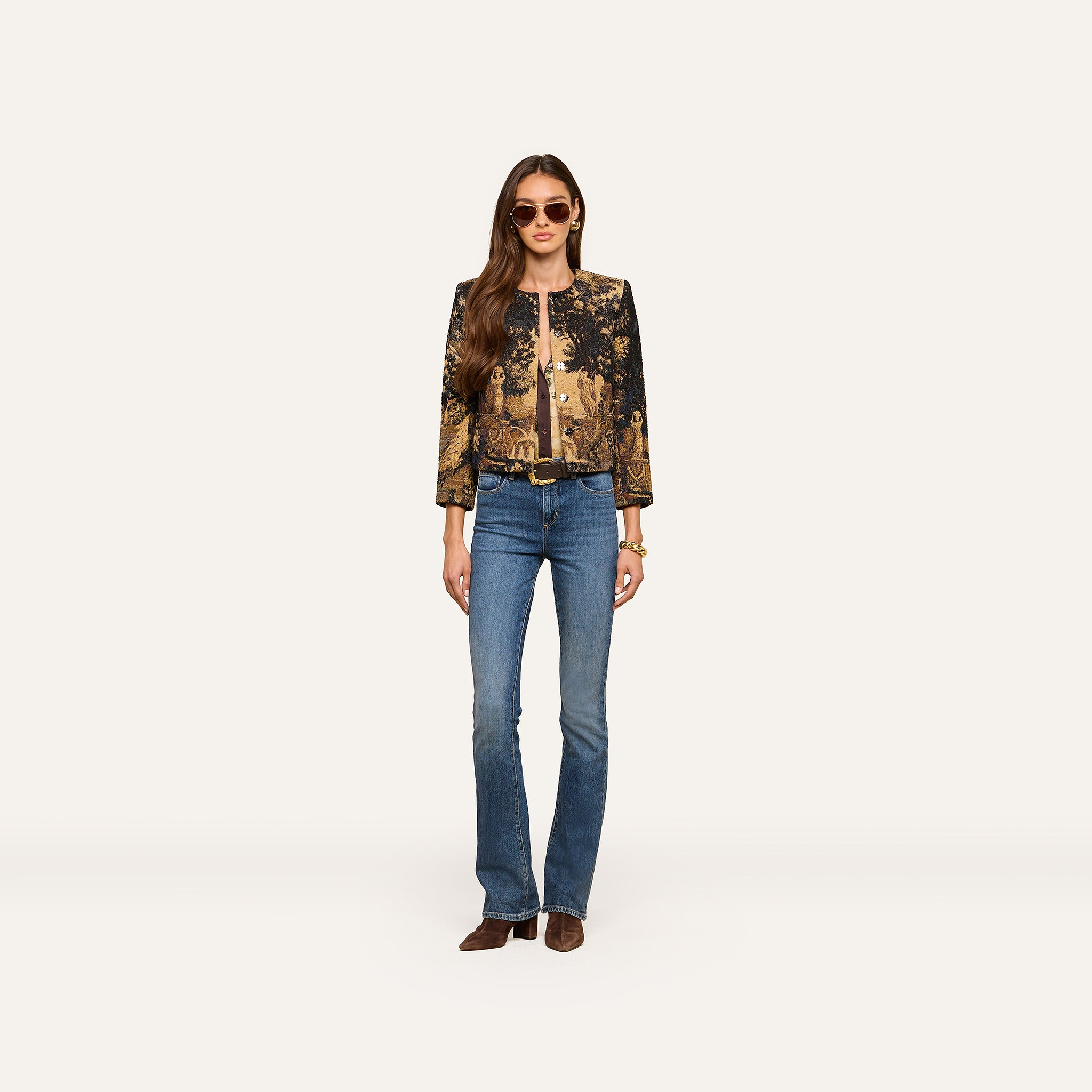 A woman stands facing forward against a plain light background, wearing sunglasses, blue jeans, brown shoes, and the Lian Tapestry Jacket, her hands by her sides.