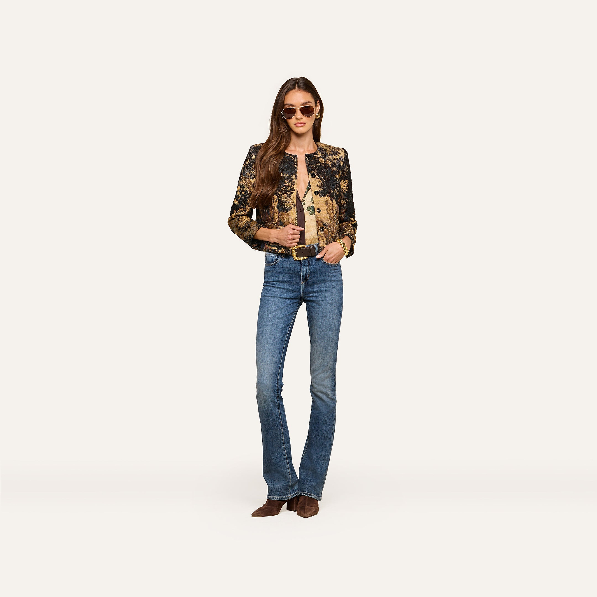 Wearing sunglasses, a person stands against a plain white background dressed in the Lian Tapestry Jacket, paired with a light brown top, blue jeans, and brown shoes.