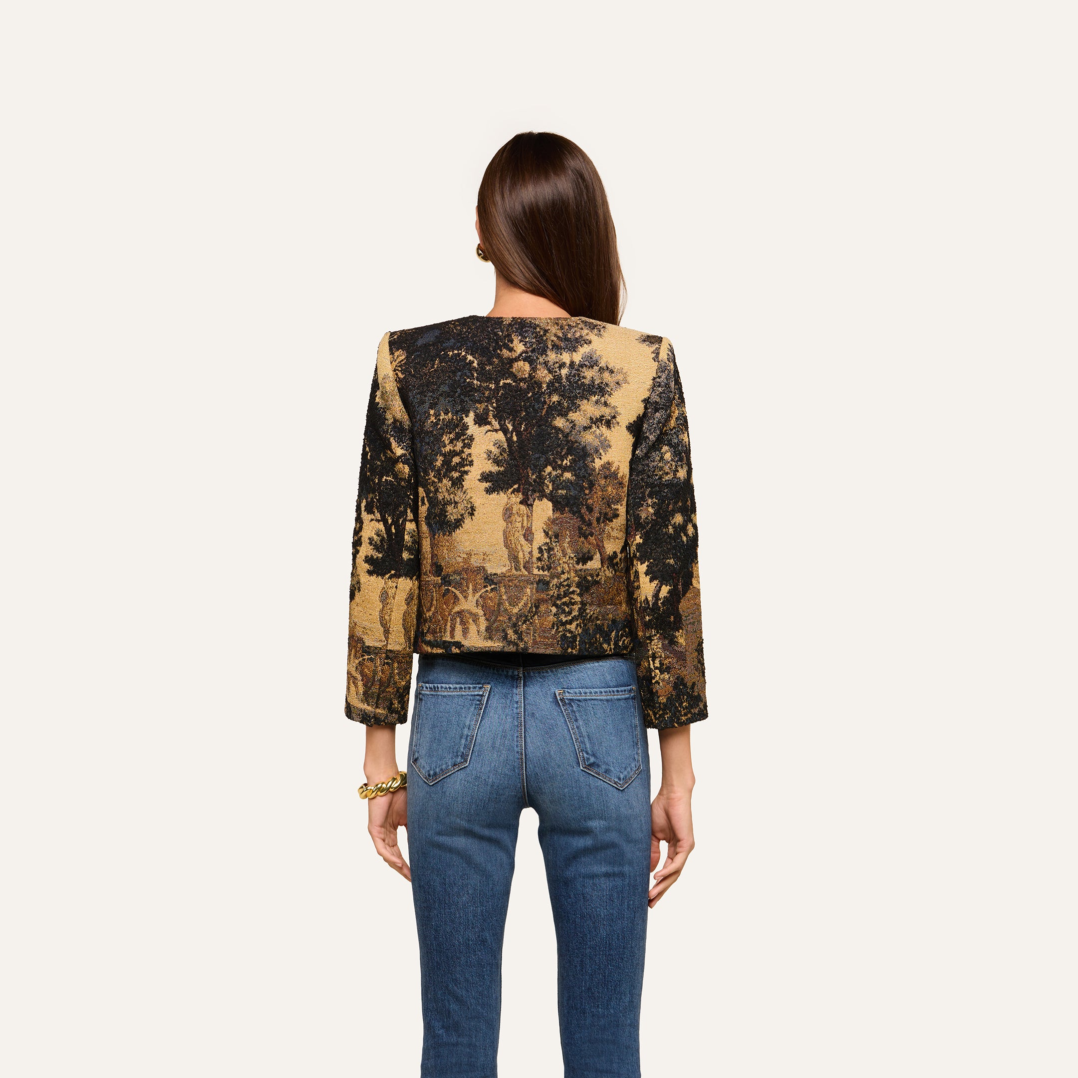 A woman with long brown hair, seen from the back, wears blue jeans and the Lian Tapestry Jacket—a cropped piece featuring a detailed landscape tapestry print inspired by Flemish designs with trees and figures.
