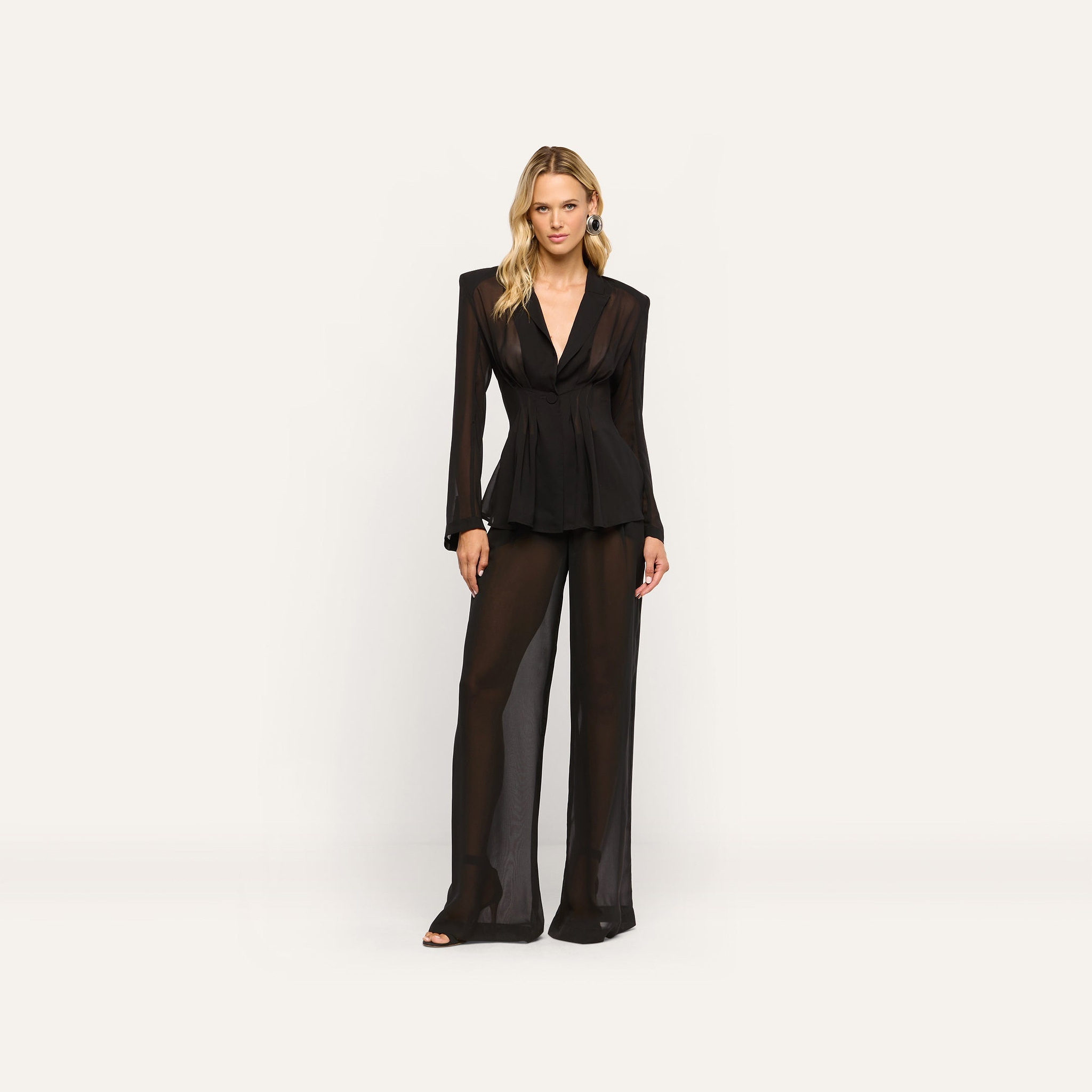 A woman wears a black sheer long-sleeved Rouvier Blazer Top with statement shoulders and matching tailored wide-leg trousers.