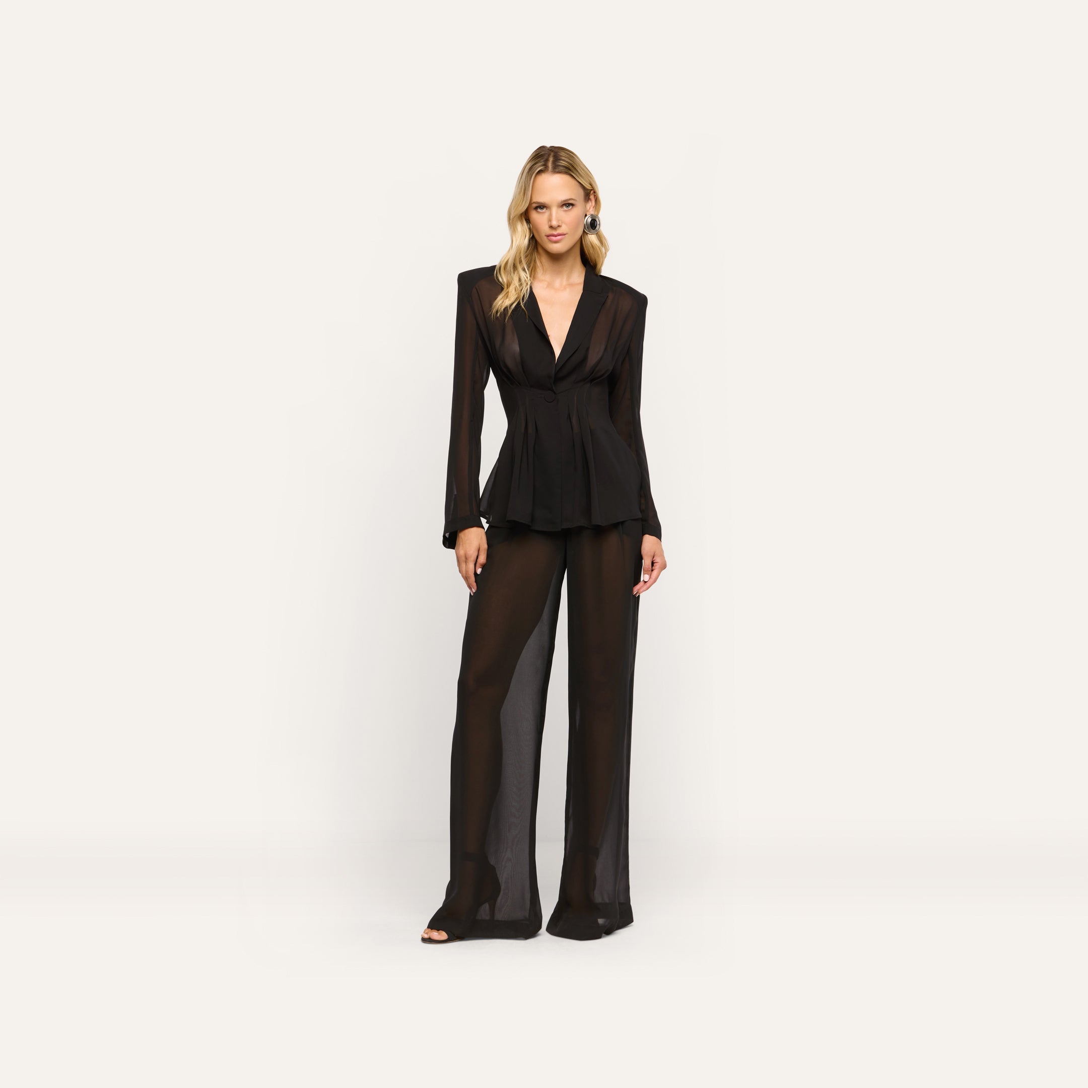 A woman wears a black sheer long-sleeved Rouvier Blazer Top with statement shoulders and matching tailored wide-leg trousers.