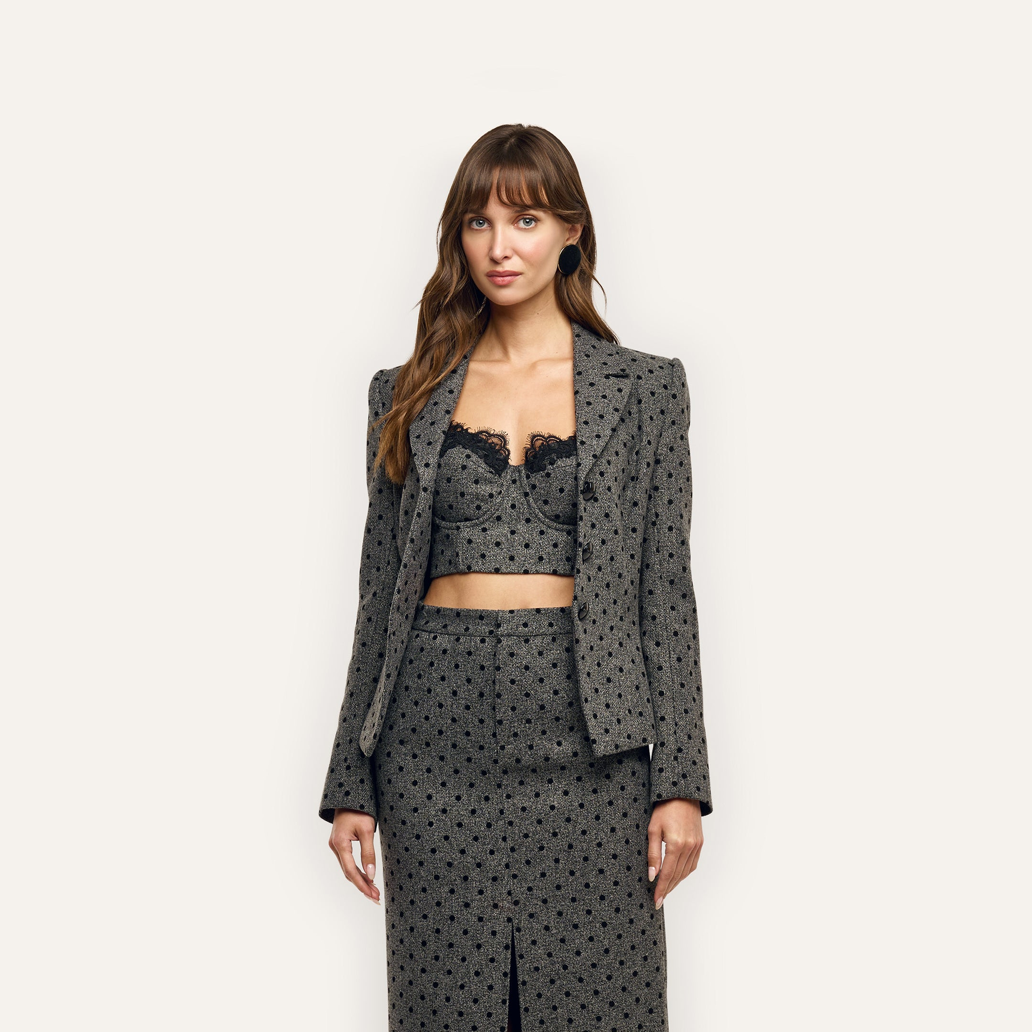 A woman wears an Odile Blazer and matching midi skirt, both tailored and patterned with black polka dots, over a black lace-trimmed bustier.
