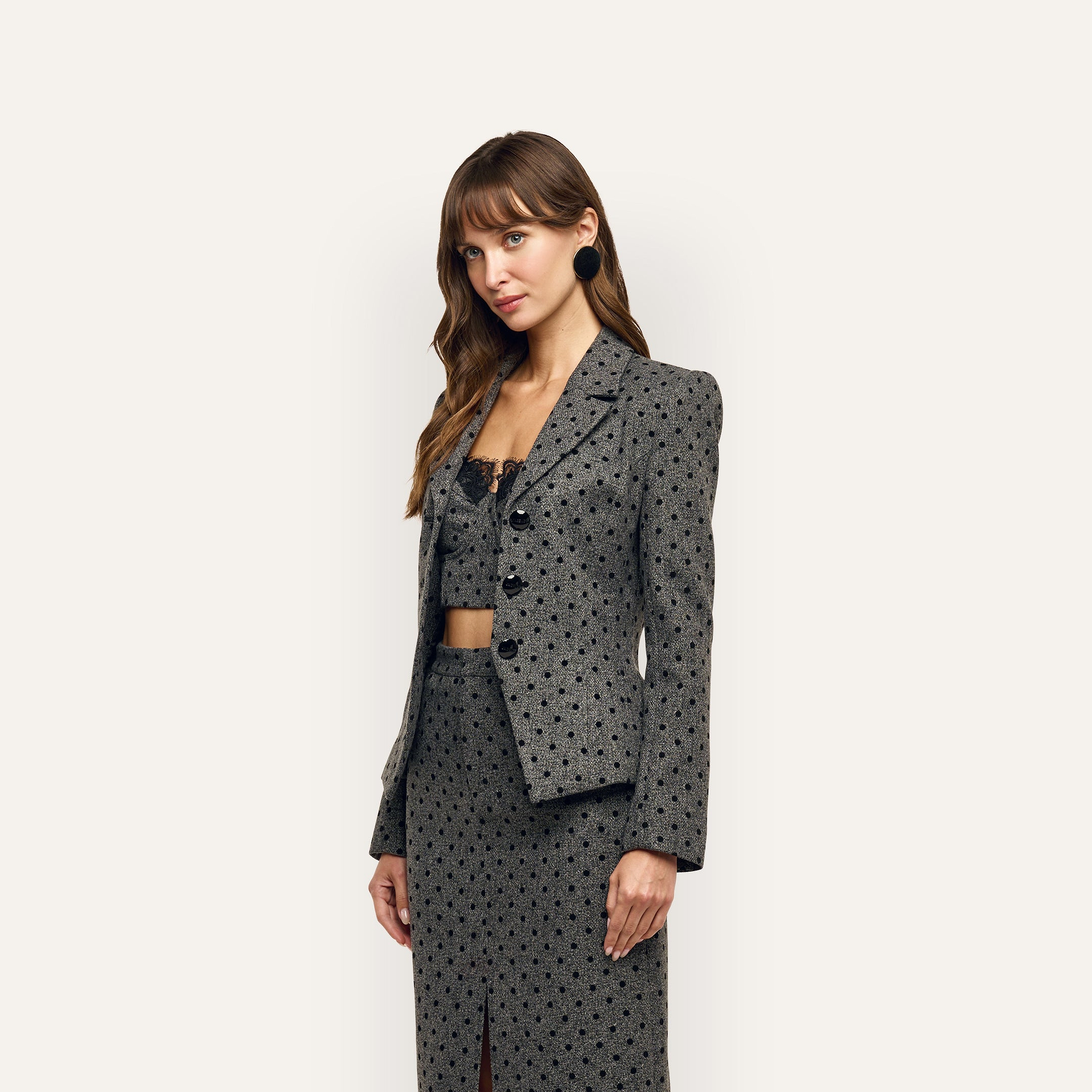 A woman wears an Odile Blazer in fitted, velvet flocked grey polka-dot over a matching cropped top and a long skirt with a front slit.