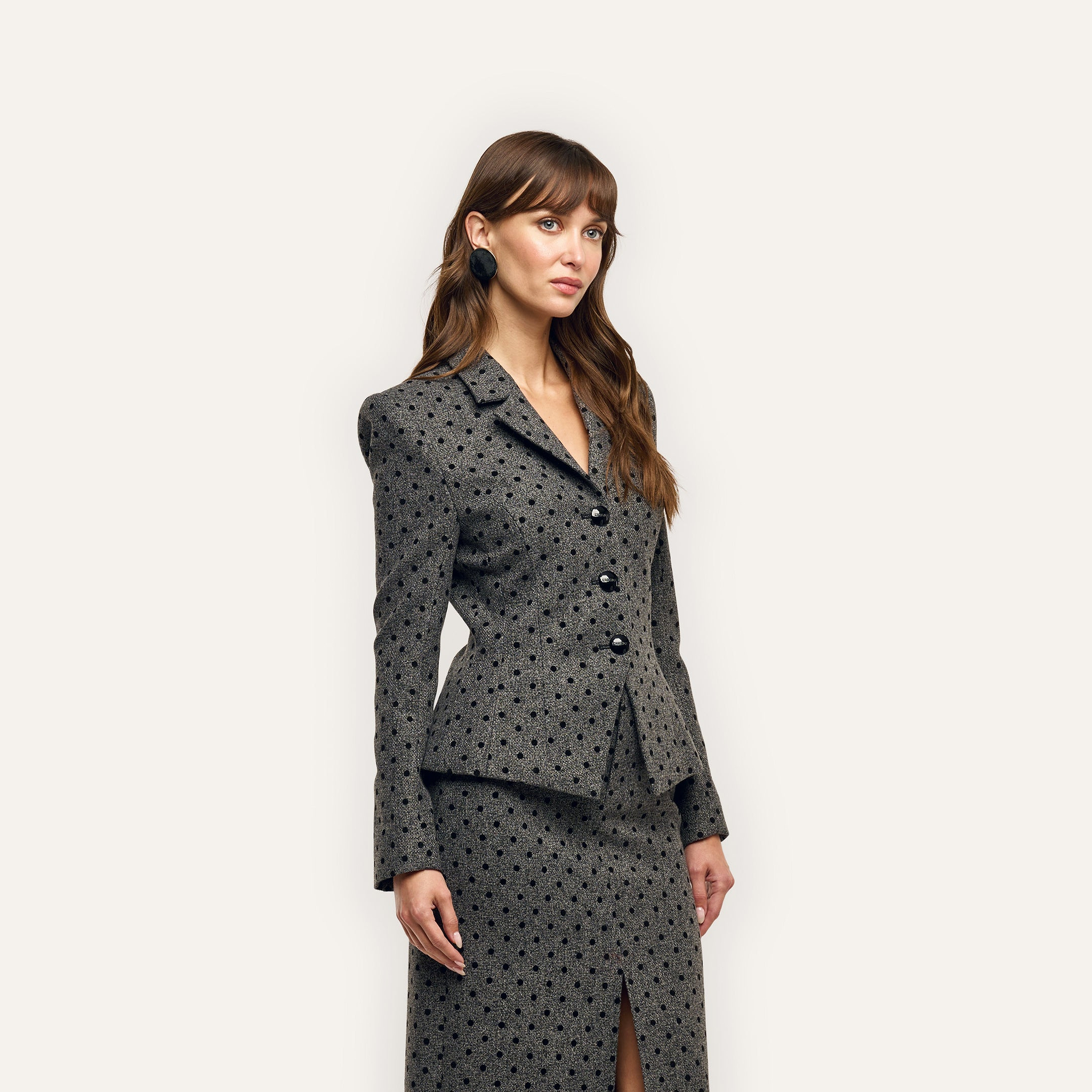 A woman wearing a charcoal-grey, velvet flocked polka-dot Odile Blazer and matching knee-length skirt with a front slit.