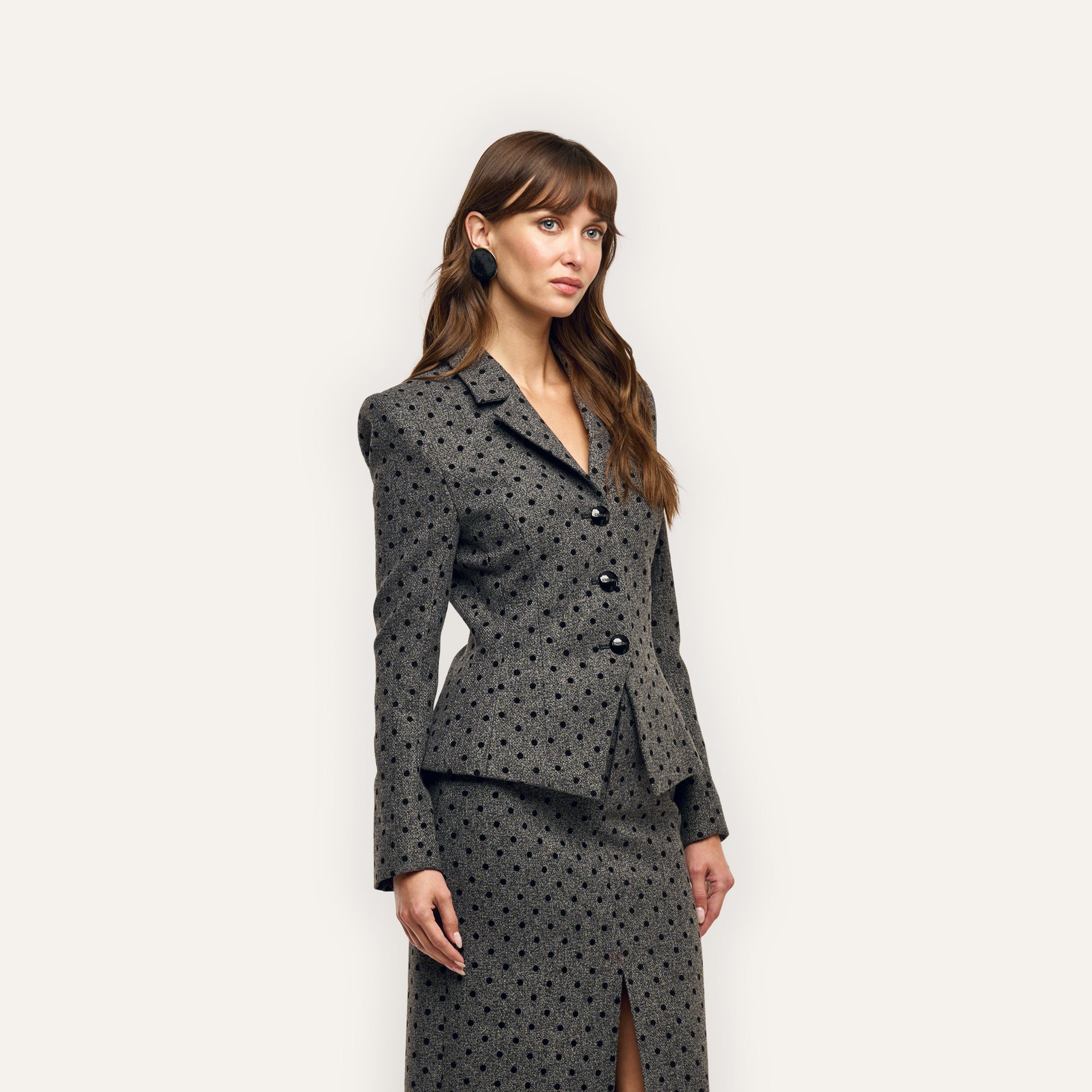 A woman wearing a charcoal-grey, velvet flocked polka-dot Odile Blazer and matching knee-length skirt with a front slit.