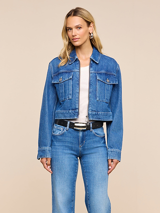 A woman wearing the Micah Oversized Jean Jacket over a white shirt and blue jeans, accessorized with a black belt, stands against a plain beige background, looking at the camera with a slight smile.
