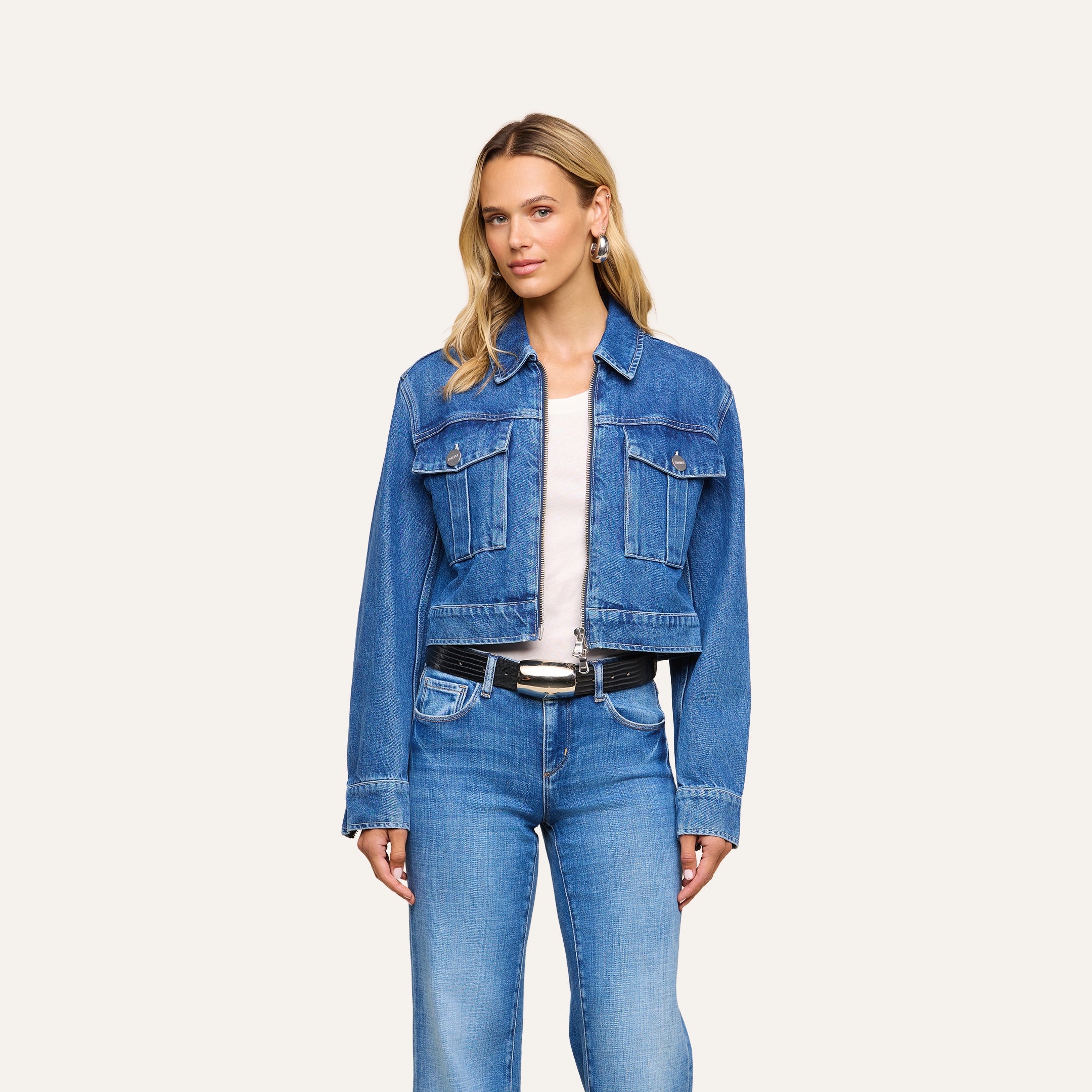 A woman with long blonde hair stands against a plain, light-colored background, wearing the Micah Oversized Jean Jacket in an indigo wash over a white shirt and blue jeans, accessorized with a black belt.