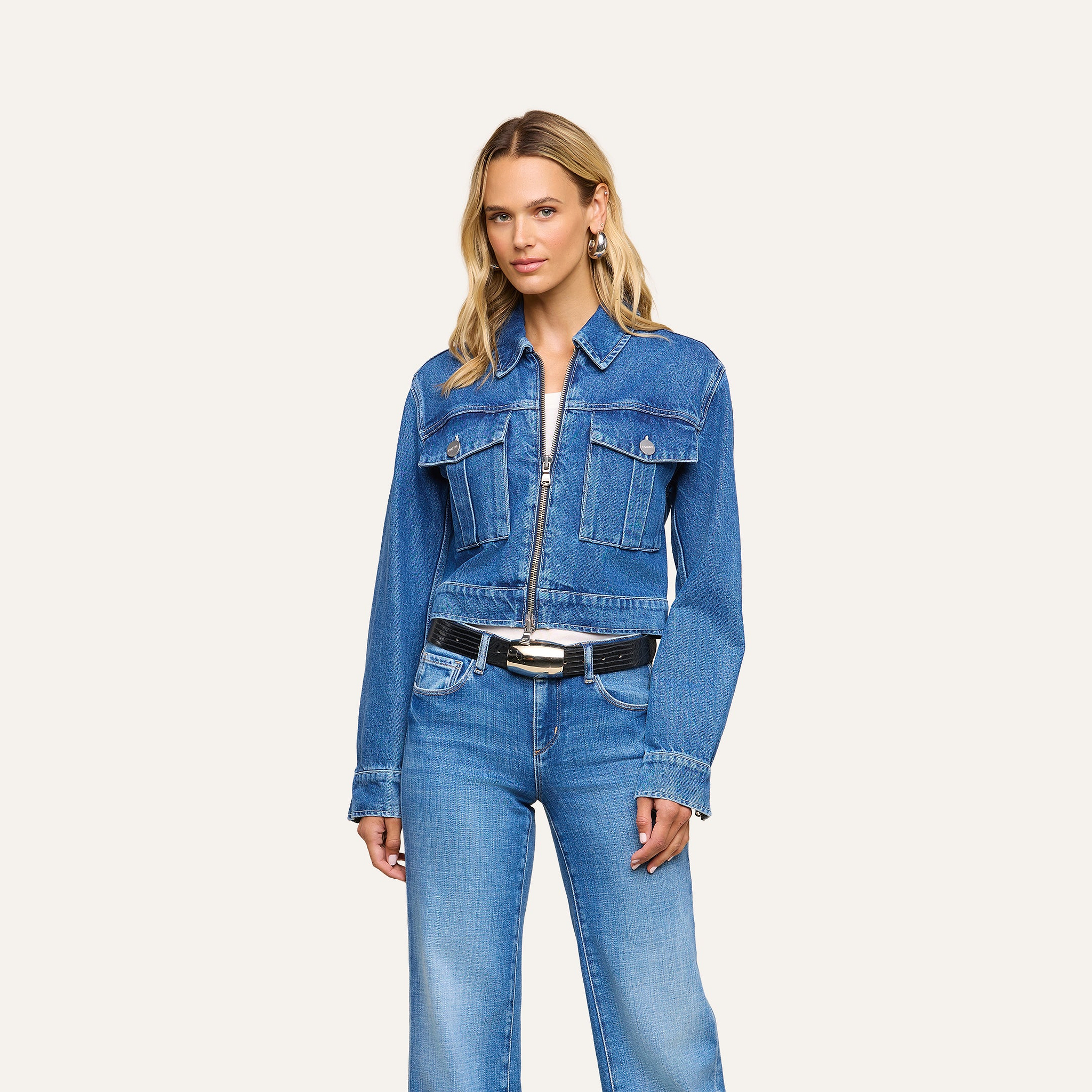A woman with long blonde hair wears the Micah Oversized Jean Jacket in an indigo wash with front pockets, paired with blue jeans, a black belt, and silver earrings, standing against a plain light background.