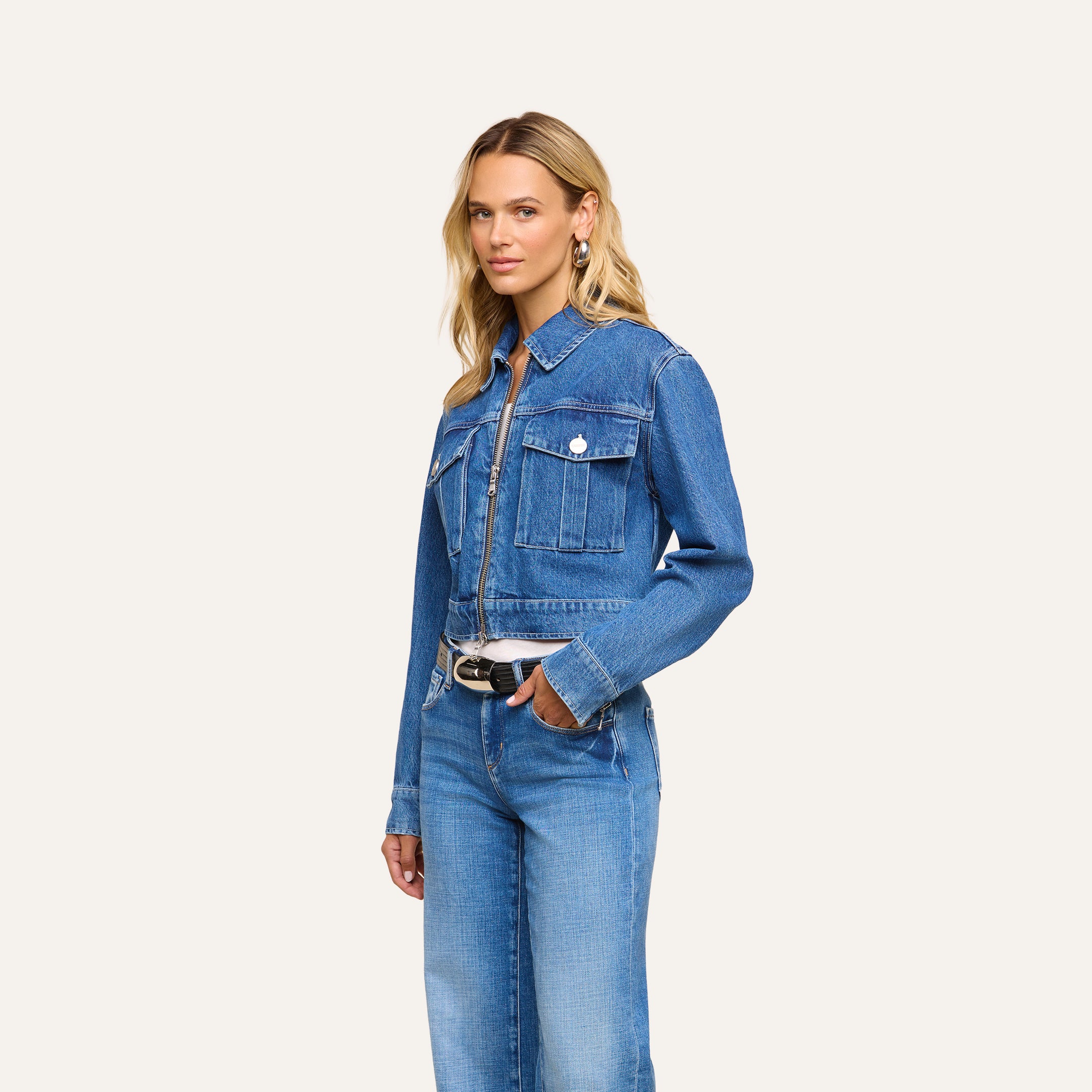 A woman with long blonde hair stands against a plain light background, looking at the camera with one hand in her pocket. She is wearing the Micah Oversized Jean Jacket in an indigo wash, paired with matching jeans.