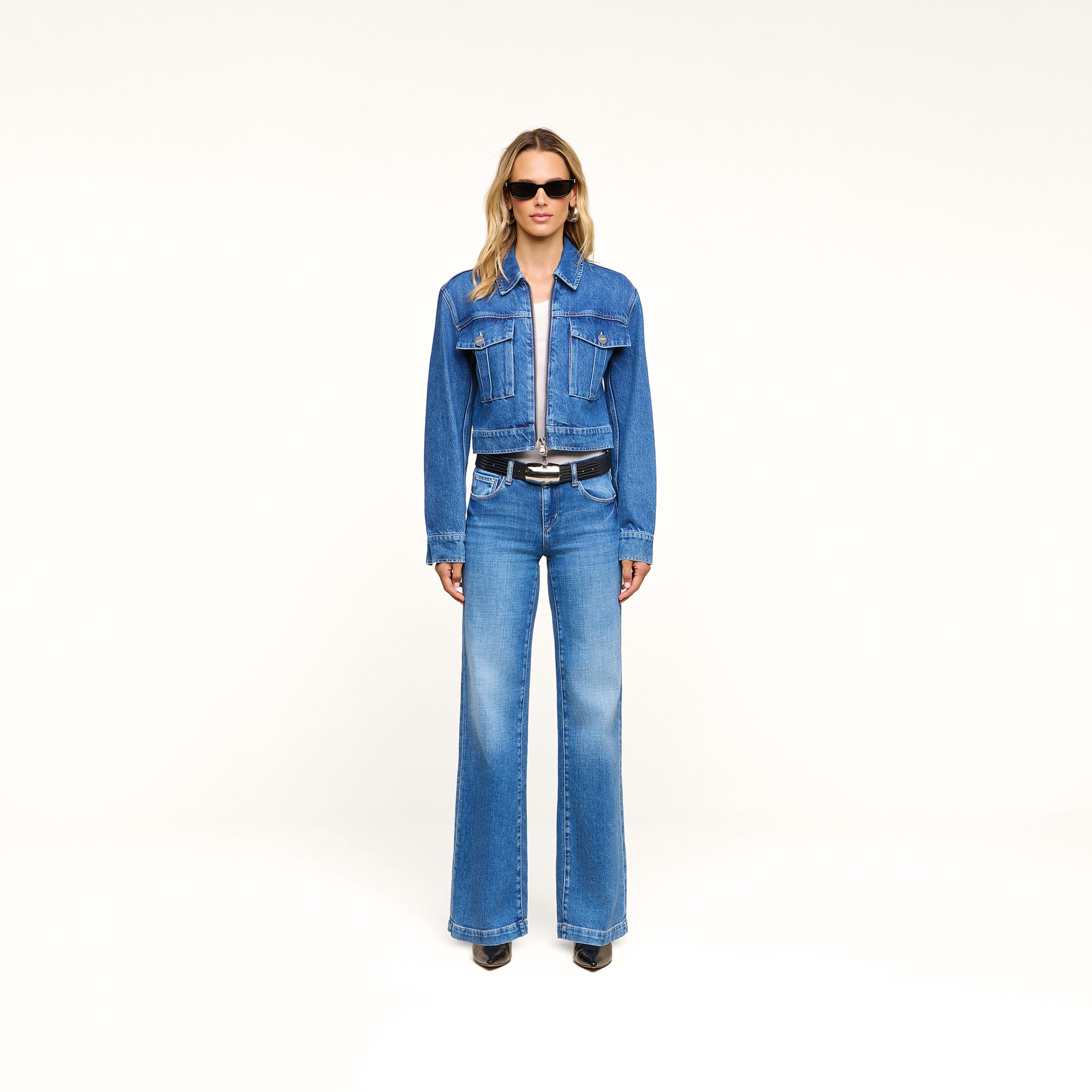 A woman stands against a white background wearing sunglasses, the Micah Oversized Jean Jacket in an indigo wash with a cropped fit, matching blue jeans, a black belt, and black shoes.