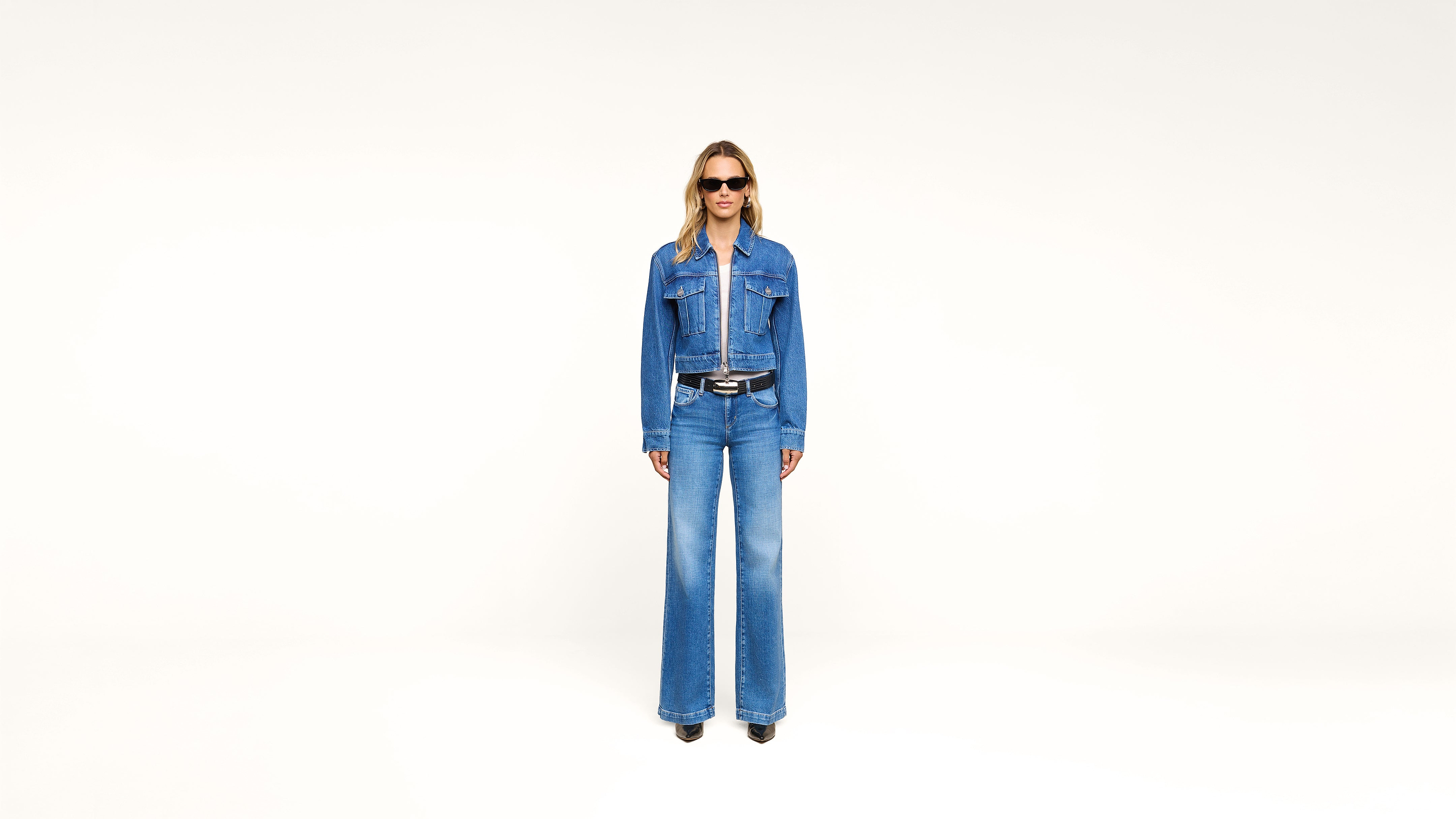 A person wearing a denim jacket and wide-leg jeans stands against a plain white background, wearing sunglasses and black shoes.