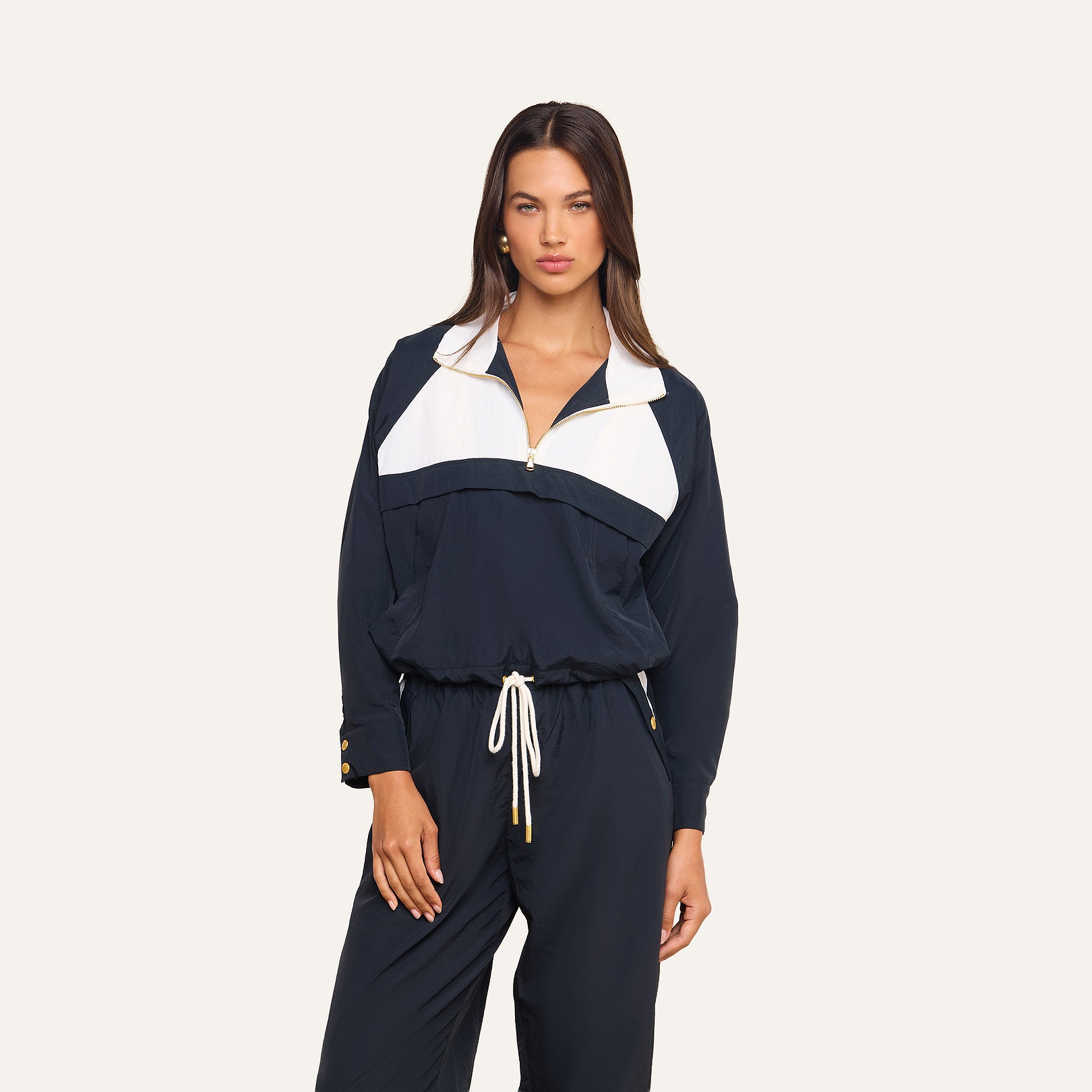 A woman wears the Vex Cropped Windbreaker in navy blue and white with matching pants, standing against a plain light background and looking at the camera with a neutral expression.