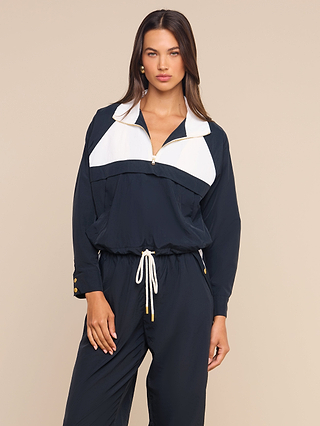 A woman stands against a beige background, looking straight ahead with one hand resting on her hip, wearing the Vex Cropped Windbreaker—a navy and white zip-up style with a drawstring waist, designed like an activewear jacket.