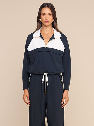 A woman wears the Vex Cropped Windbreaker, a navy and white water-repellent jacket with long sleeves, paired with matching drawstring pants. She stands against a plain beige background, gazing directly ahead with a neutral expression.